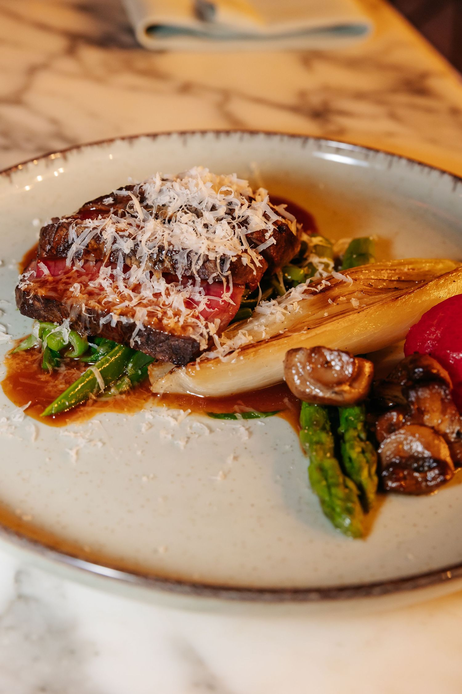 Elegant plated dish with braised meat, asparagus, mushrooms, and shaved parmesan in rich sauce on ceramic plate.
