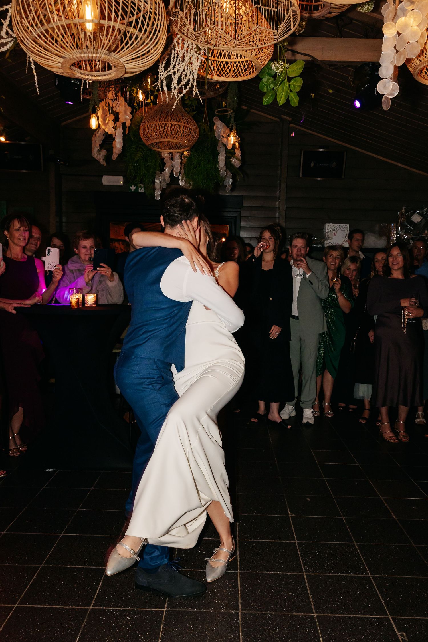Bride and groom sharing a romantic first dance embrace surrounded by wedding guests.