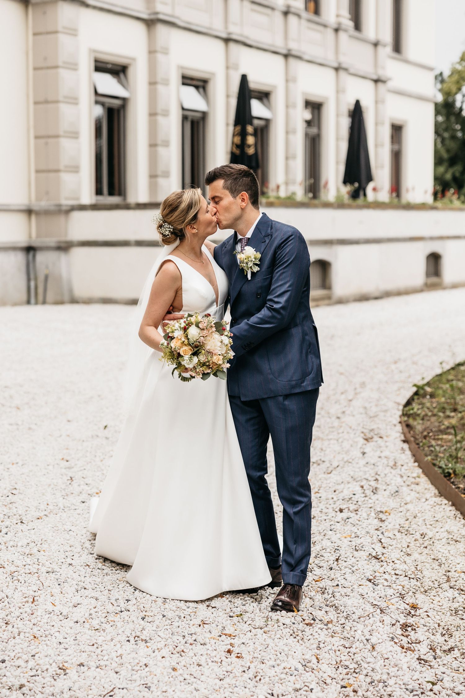 Bride and groom sharing a kiss outdoors on gravel path in front of elegant building, bride holding pastel bouquet.