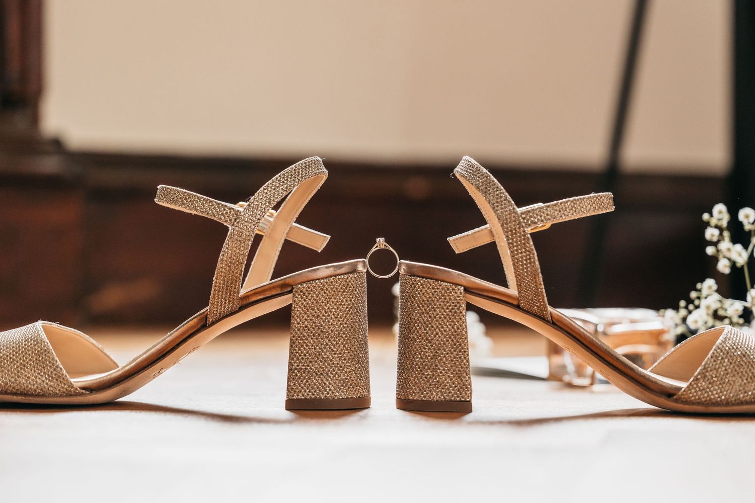 Wedding ring balanced between two gold block-heel sandals with baby's breath flowers in background.