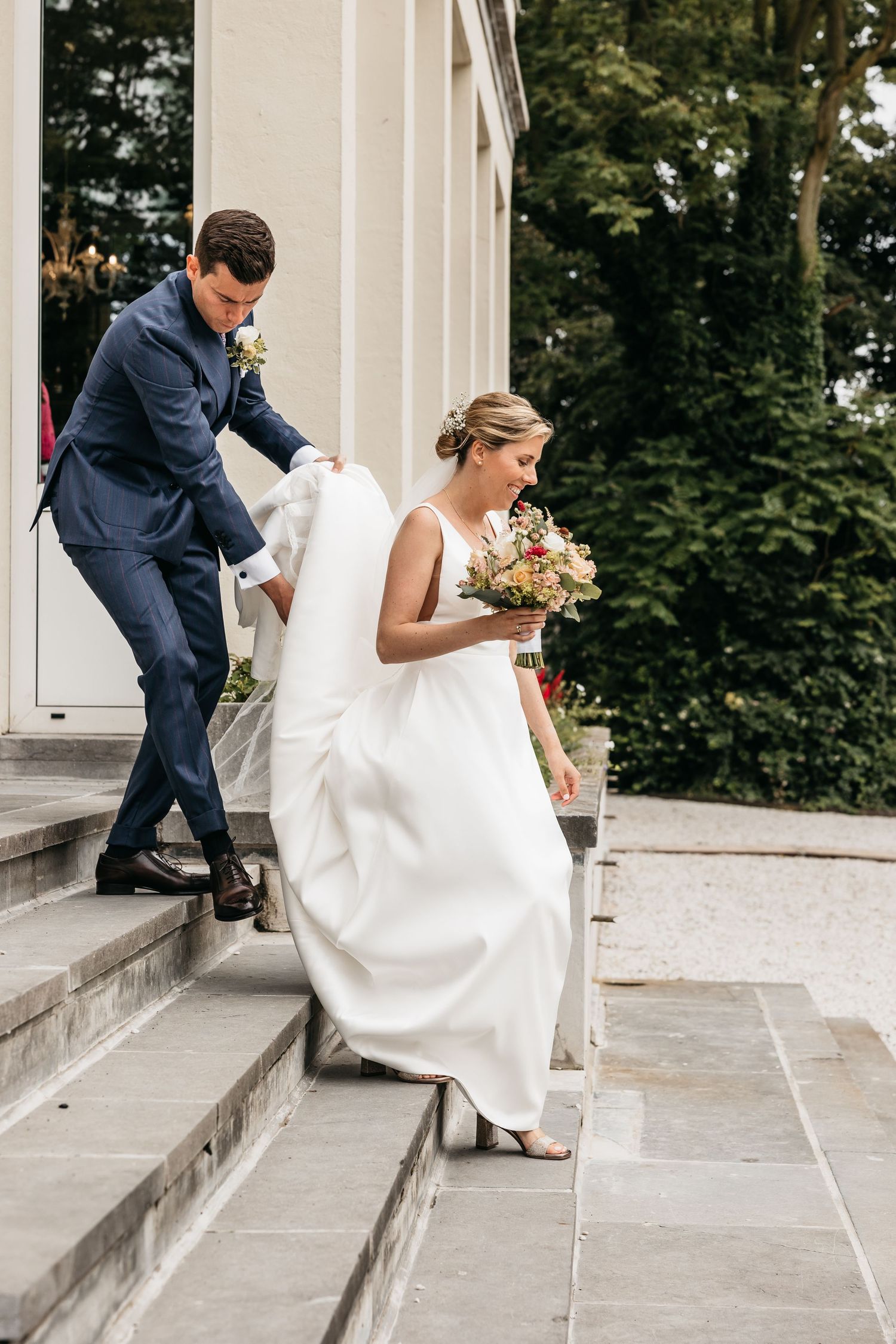 Groom in navy suit helps bride in white gown descend stone steps while she holds a floral bouquet.