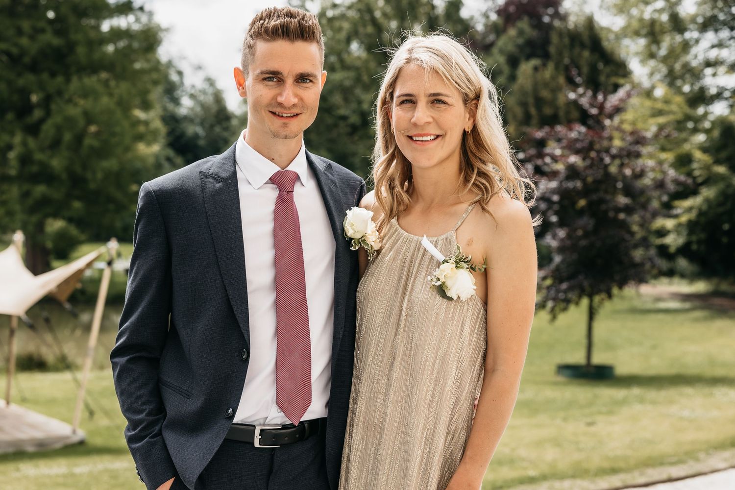 Smiling couple at a wedding, man in navy suit with pink tie and woman in a gold dress with white corsage.