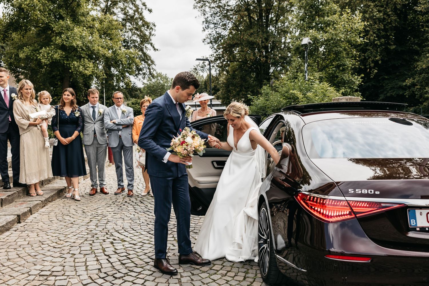 Groom helping bride in white gown exit a black Mercedes S580 luxury wedding car outdoors.