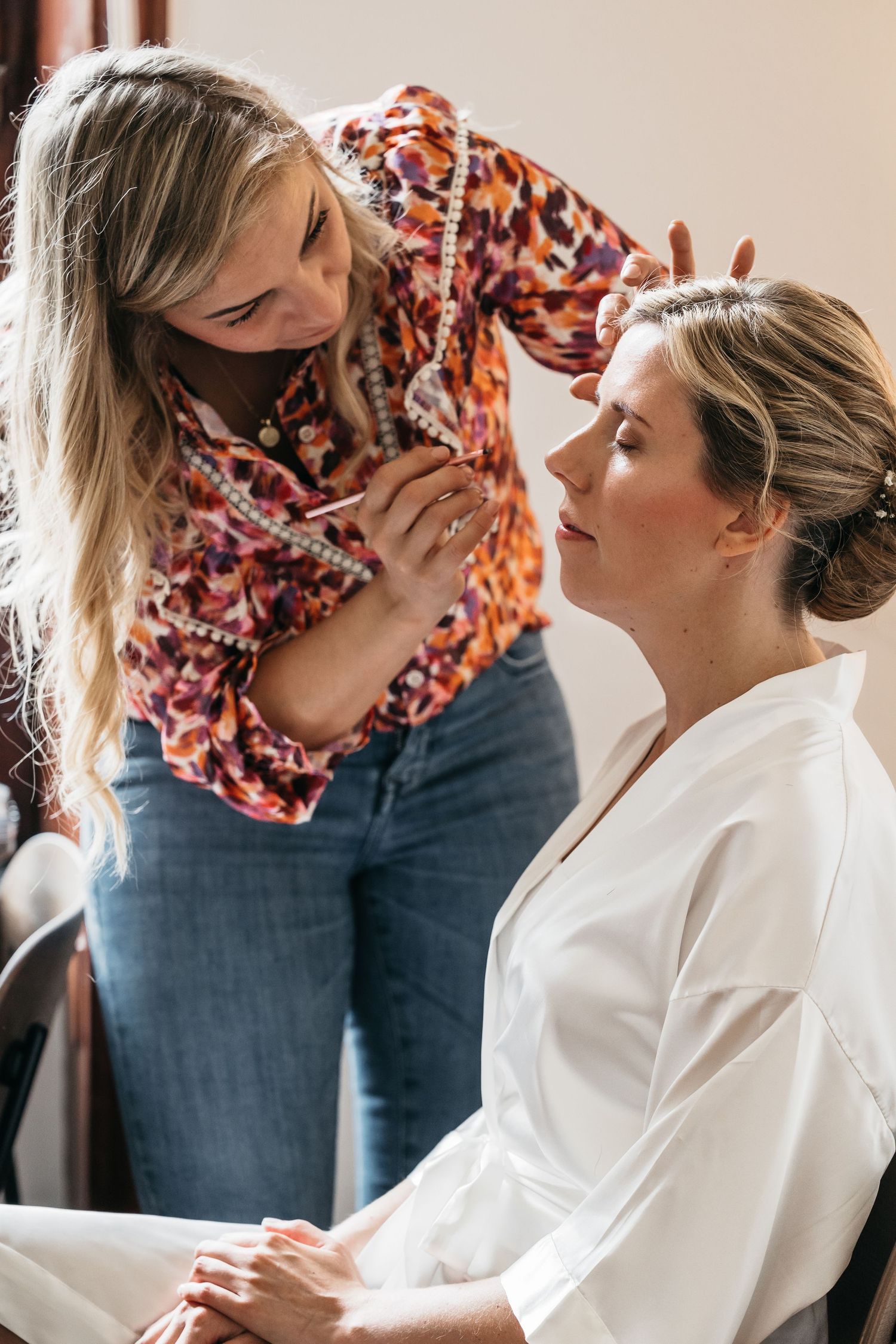 Makeup artist applying makeup to a bride in a white robe before wedding.