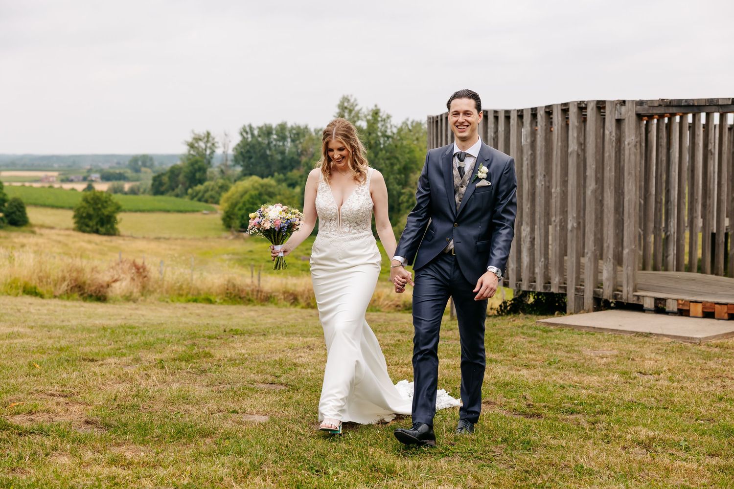 Smiling newlyweds holding hands and walking through a lush green meadow on their wedding day.