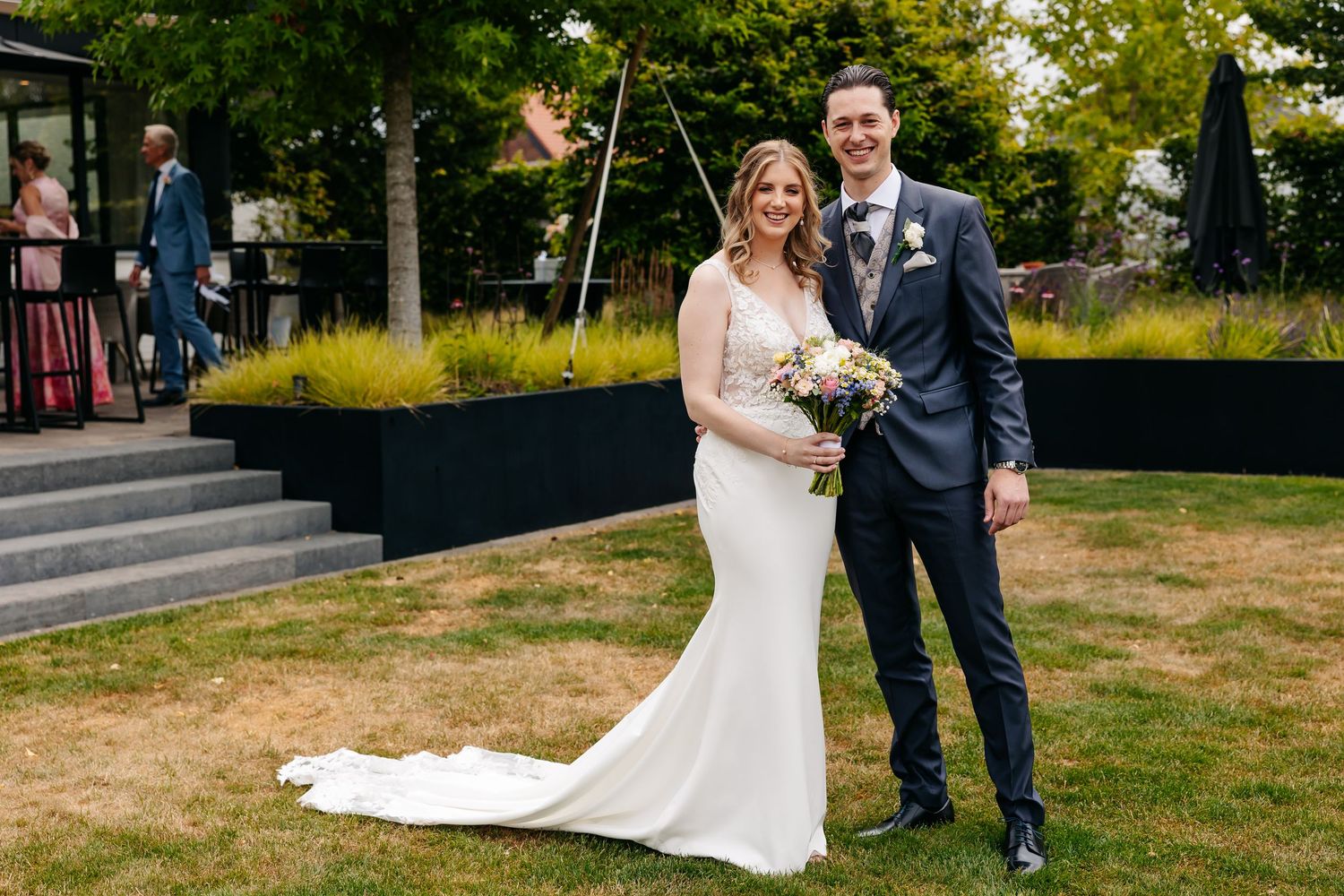 Happy bride and groom pose for formal wedding portrait on green lawn with modern venue building in background.