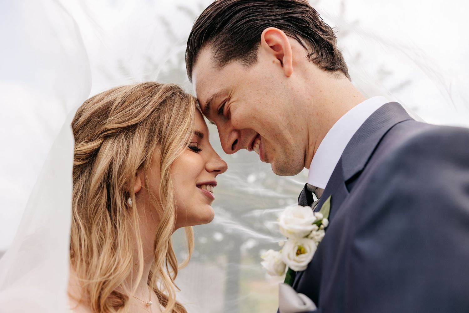 Bride and groom touch foreheads tenderly under a veil in an intimate romantic wedding portrait.
