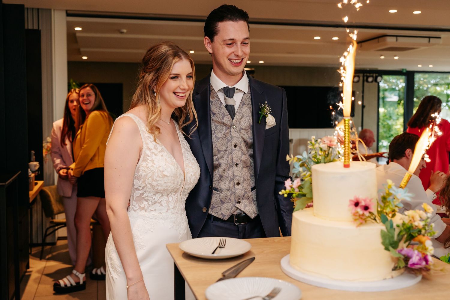 Smiling bride and groom stand beside white wedding cake with sparklers.