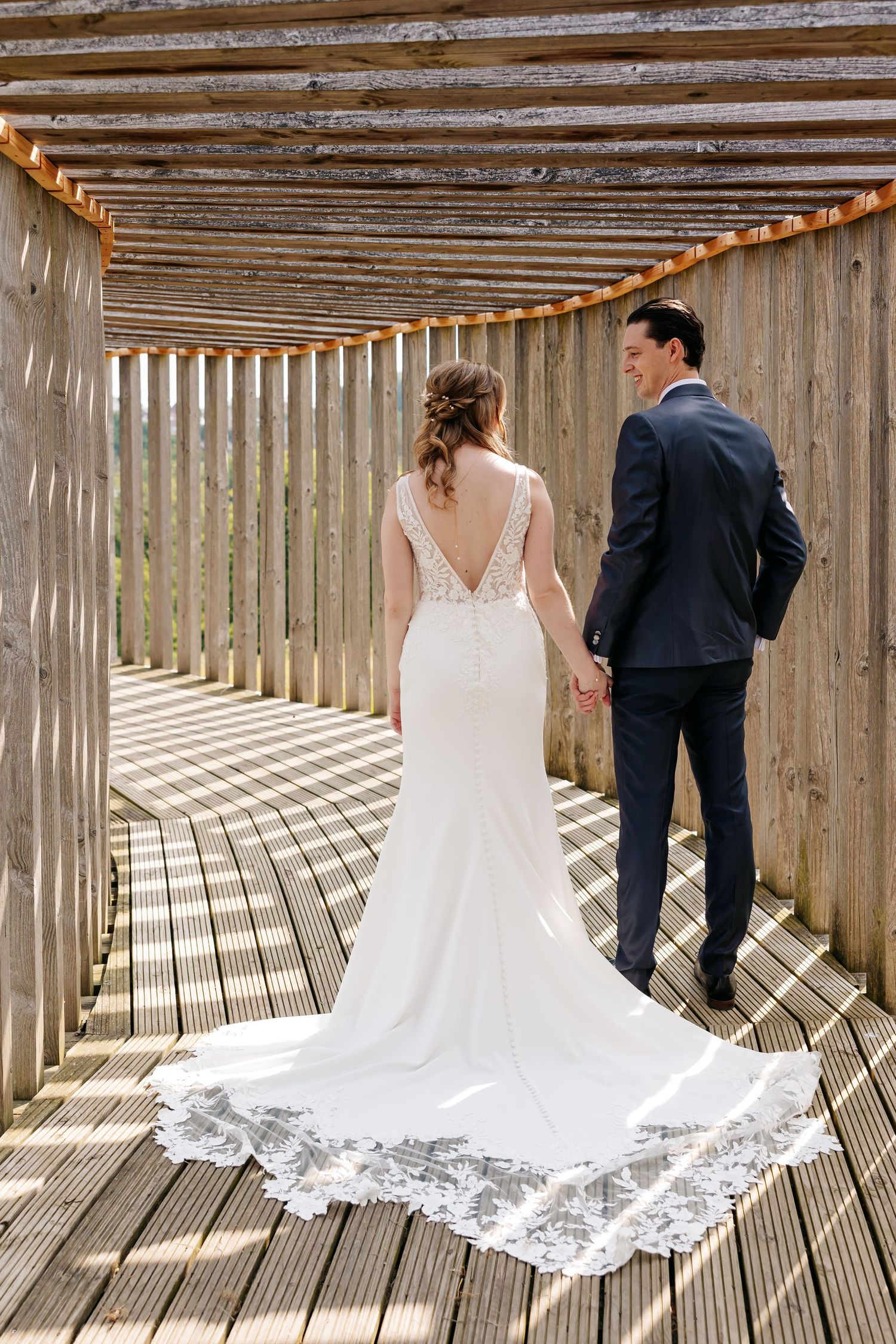 Bride and groom holding hands walking away through a rustic wooden pergola, showing lace dress train.