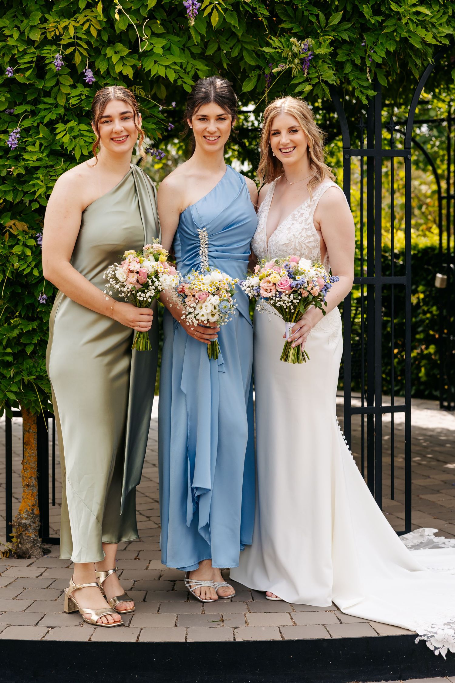 Bride in white lace gown with two bridesmaids in sage and blue dresses holding floral bouquets outdoors.