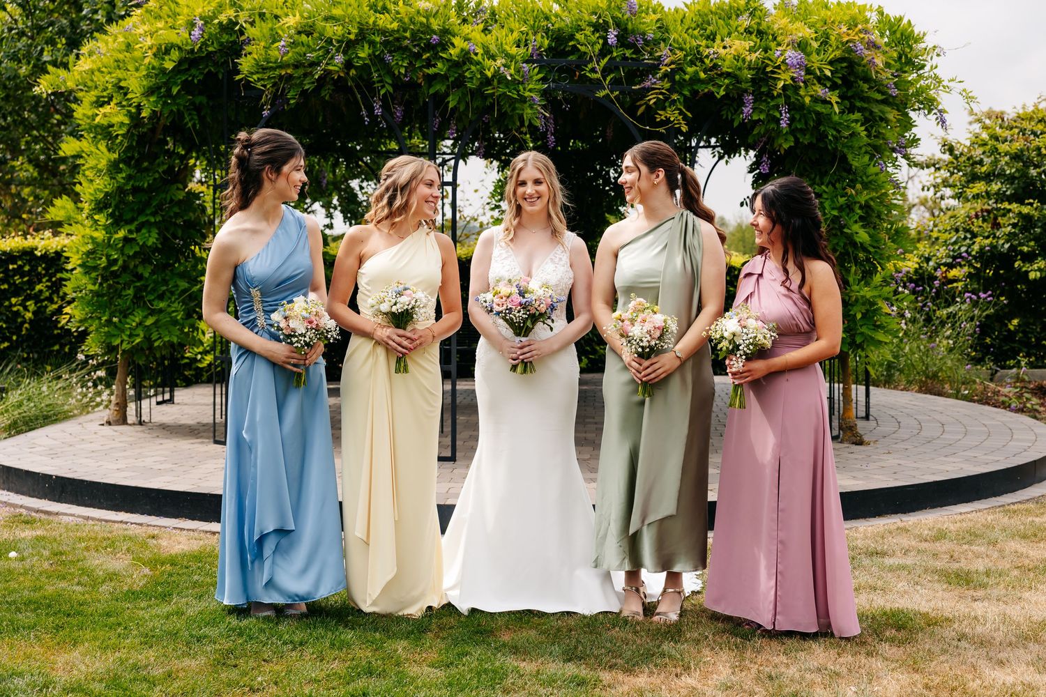 Bride in white gown poses with four bridesmaids in pastel blue, yellow, green, and pink dresses in a garden.