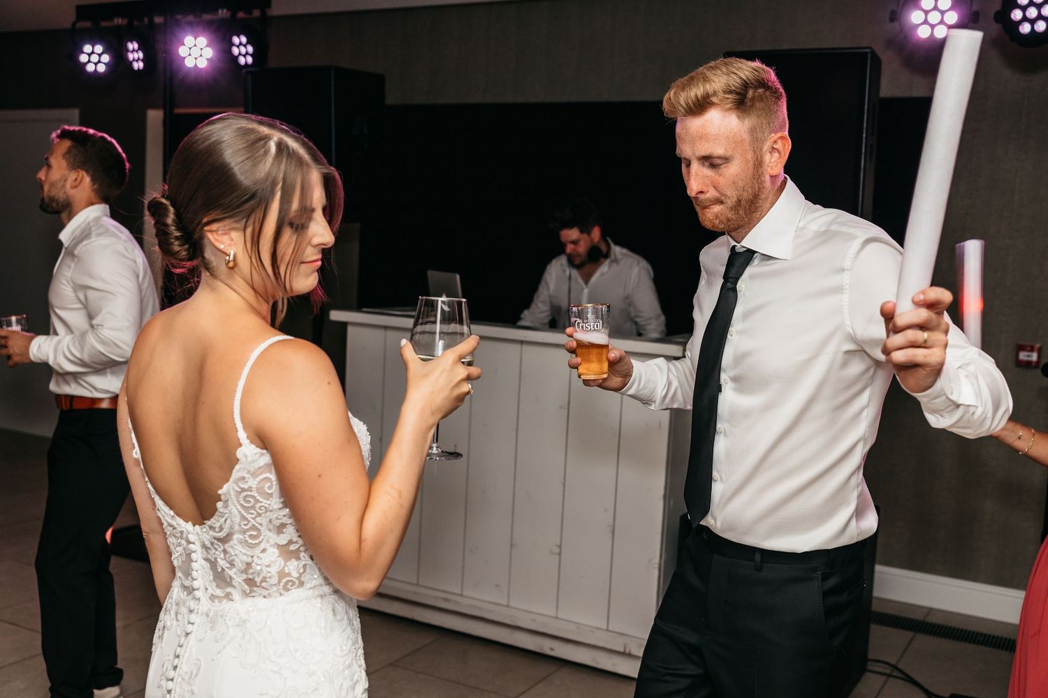Bride in backless lace gown and groomsman enjoy drinks together at wedding reception party.