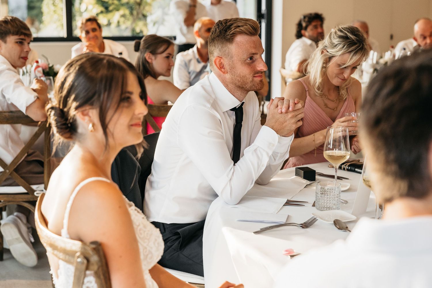Bride and groom sitting at wedding reception table smiling during speeches.