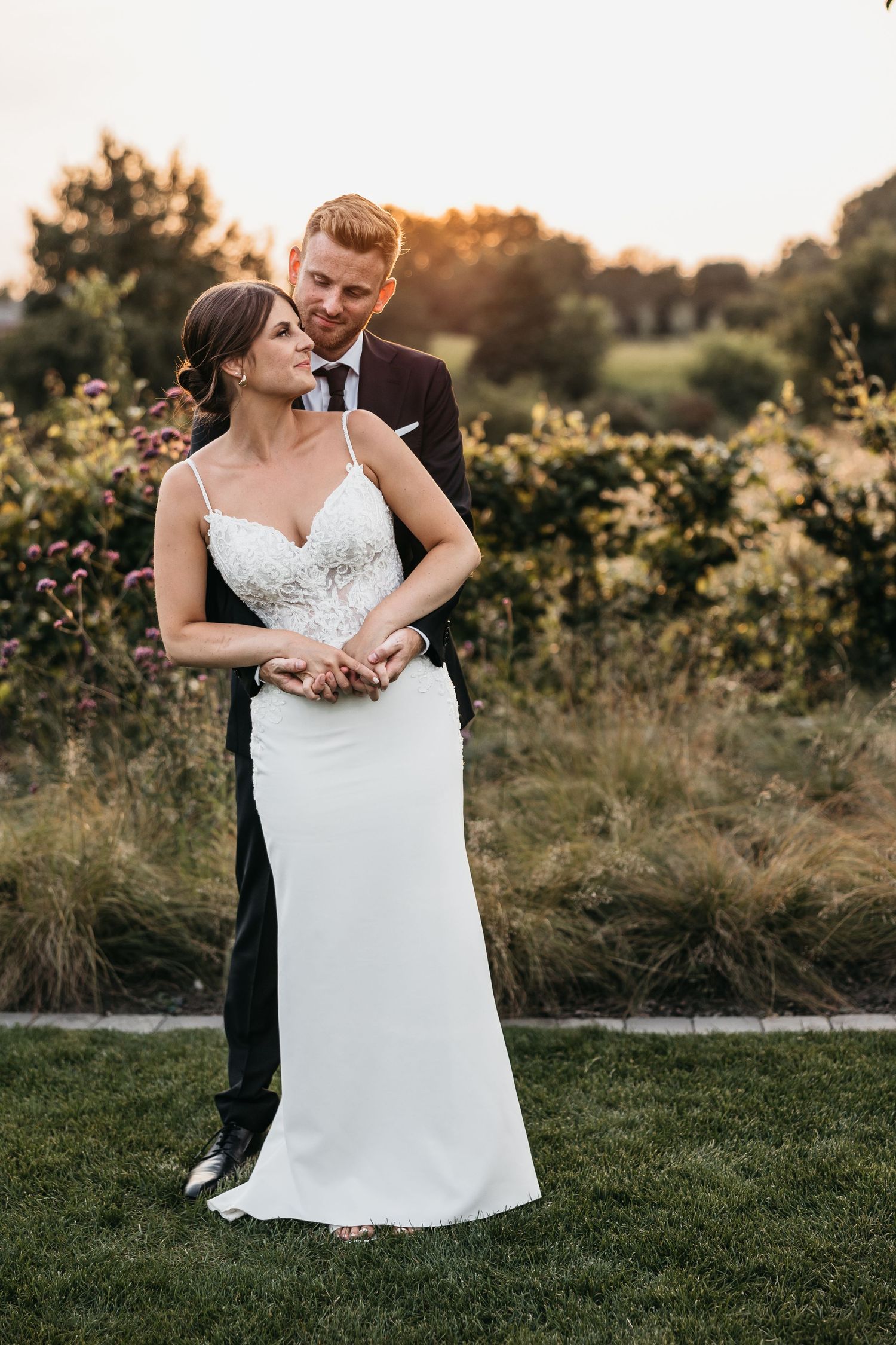 Groom in black suit embraces bride from behind in white lace gown, posing in a lush garden at golden hour sunset.