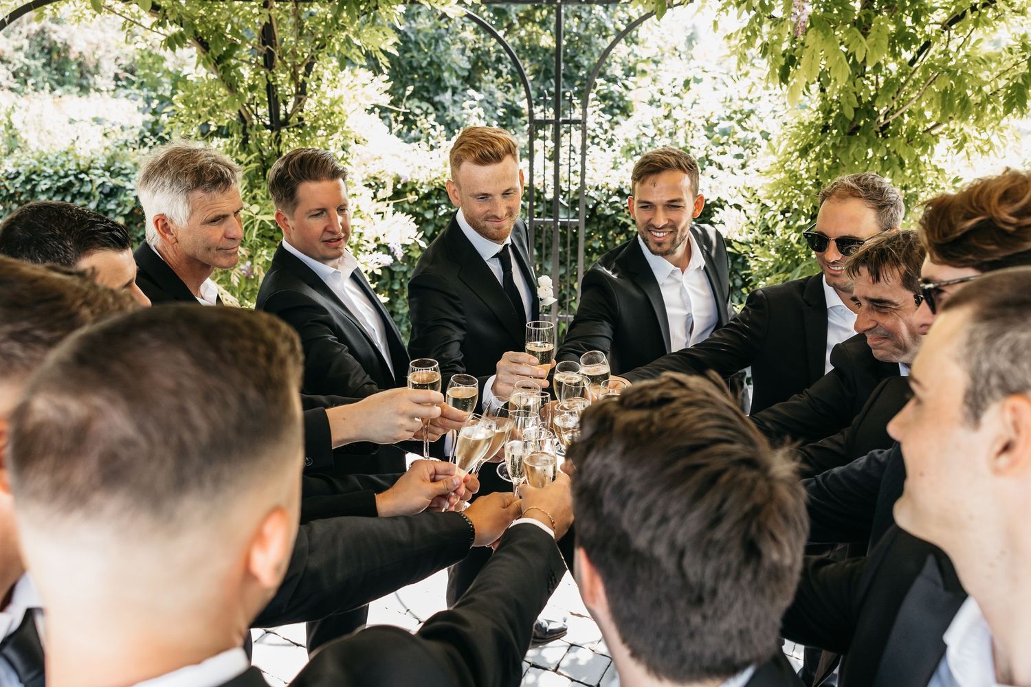 Groomsmen in black suits toasting with champagne glasses outdoors under green foliage.