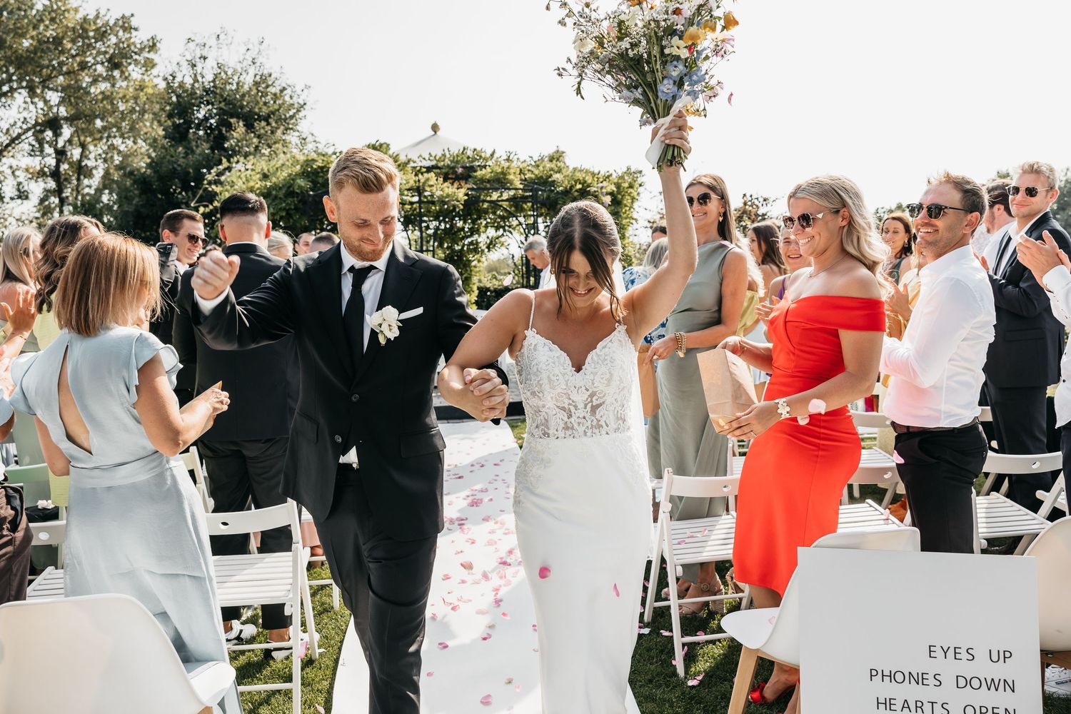 Joyful bride and groom walk down the aisle holding hands at sunny outdoor wedding ceremony.