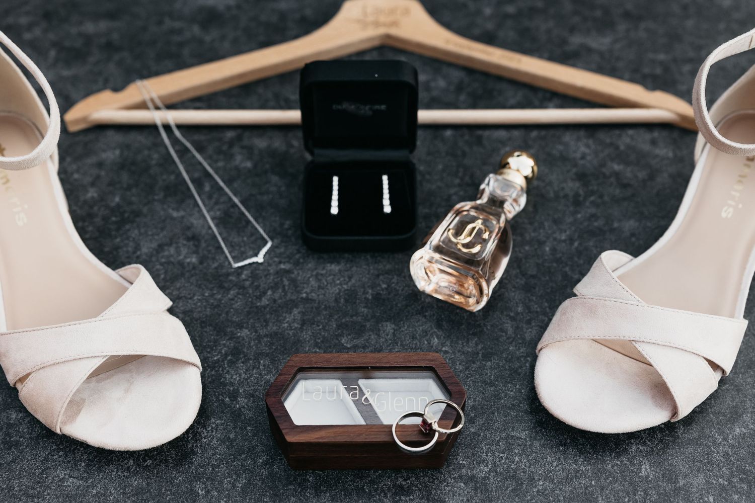 Bridal flat lay featuring white heels, diamond earrings, perfume, wedding rings, and personalized hanger.