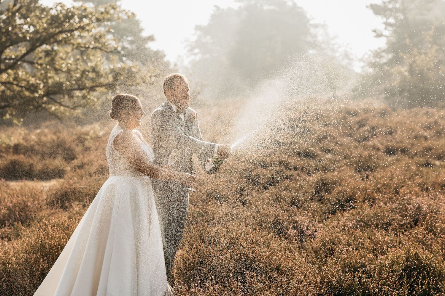 Groom spraying champagne bottle as bride laughs in sunlit heather field during wedding shoot.