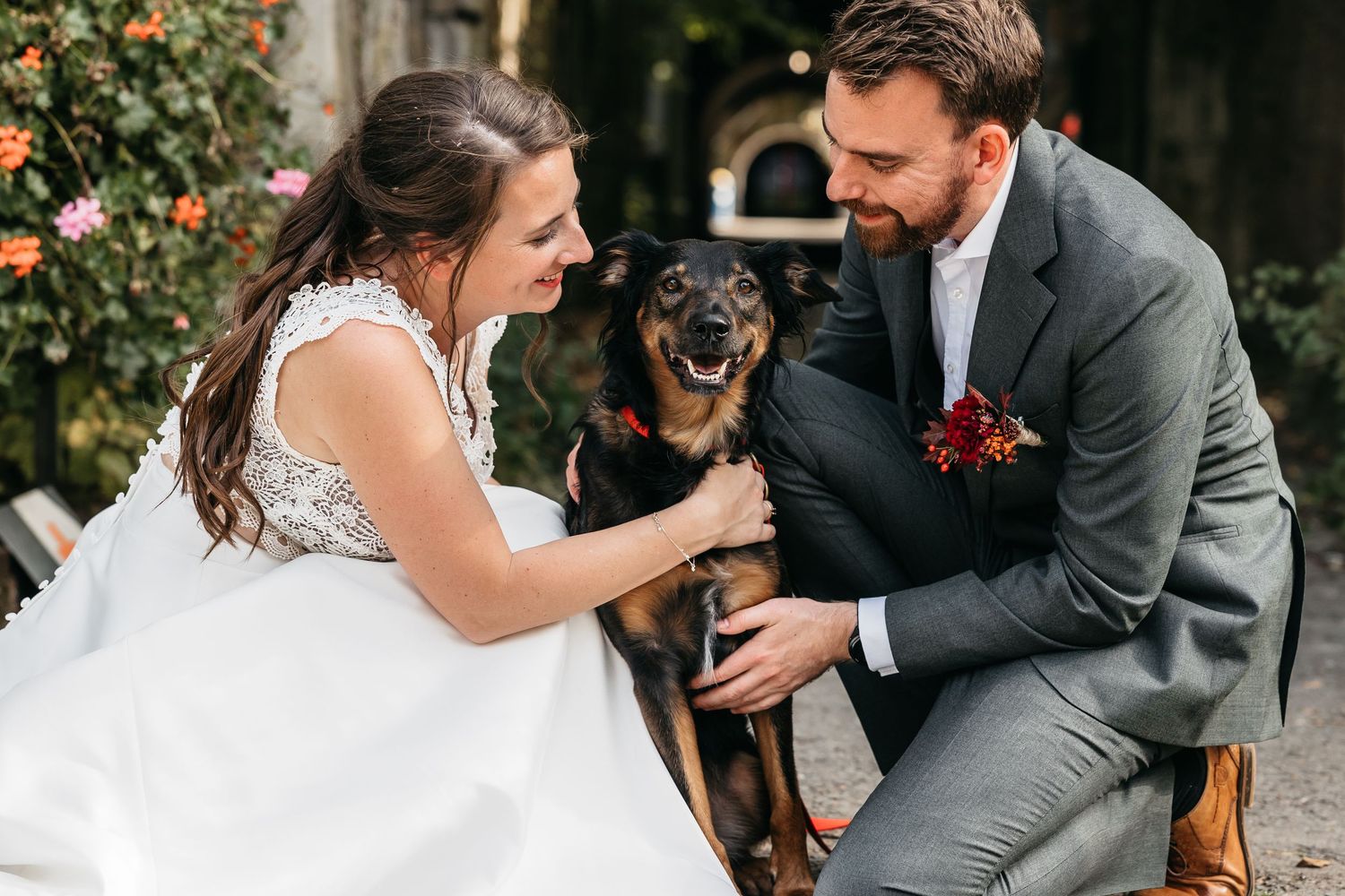 Happy bride and groom in wedding attire petting their black and tan dog outdoors.