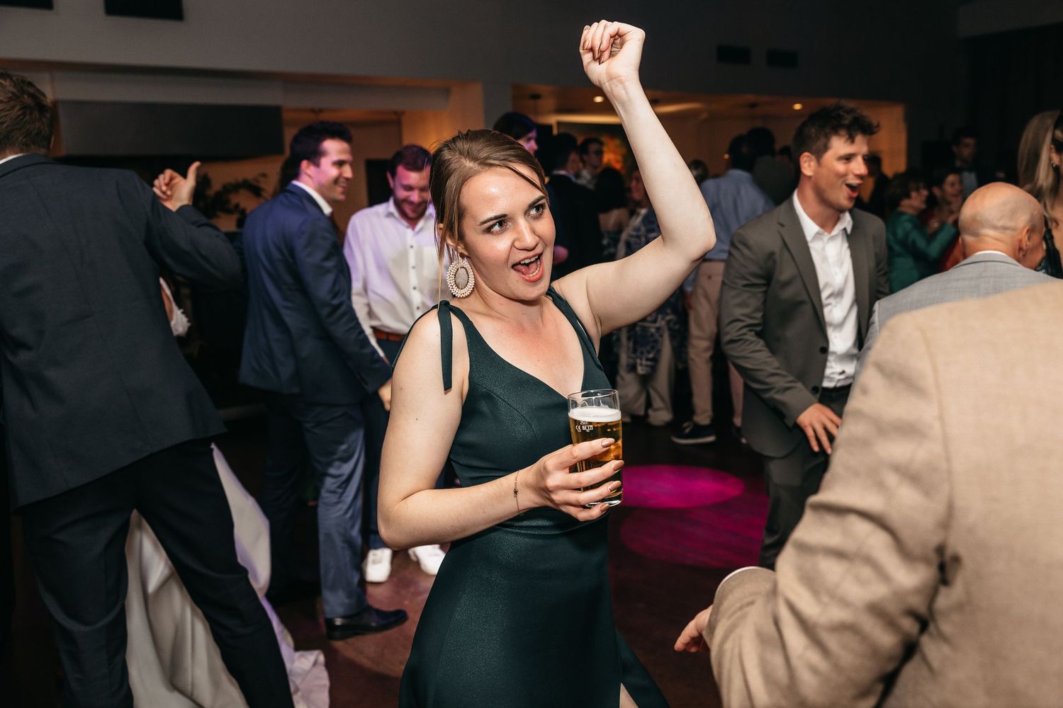 Excited woman in green dress raises arm while holding beer at wedding reception.