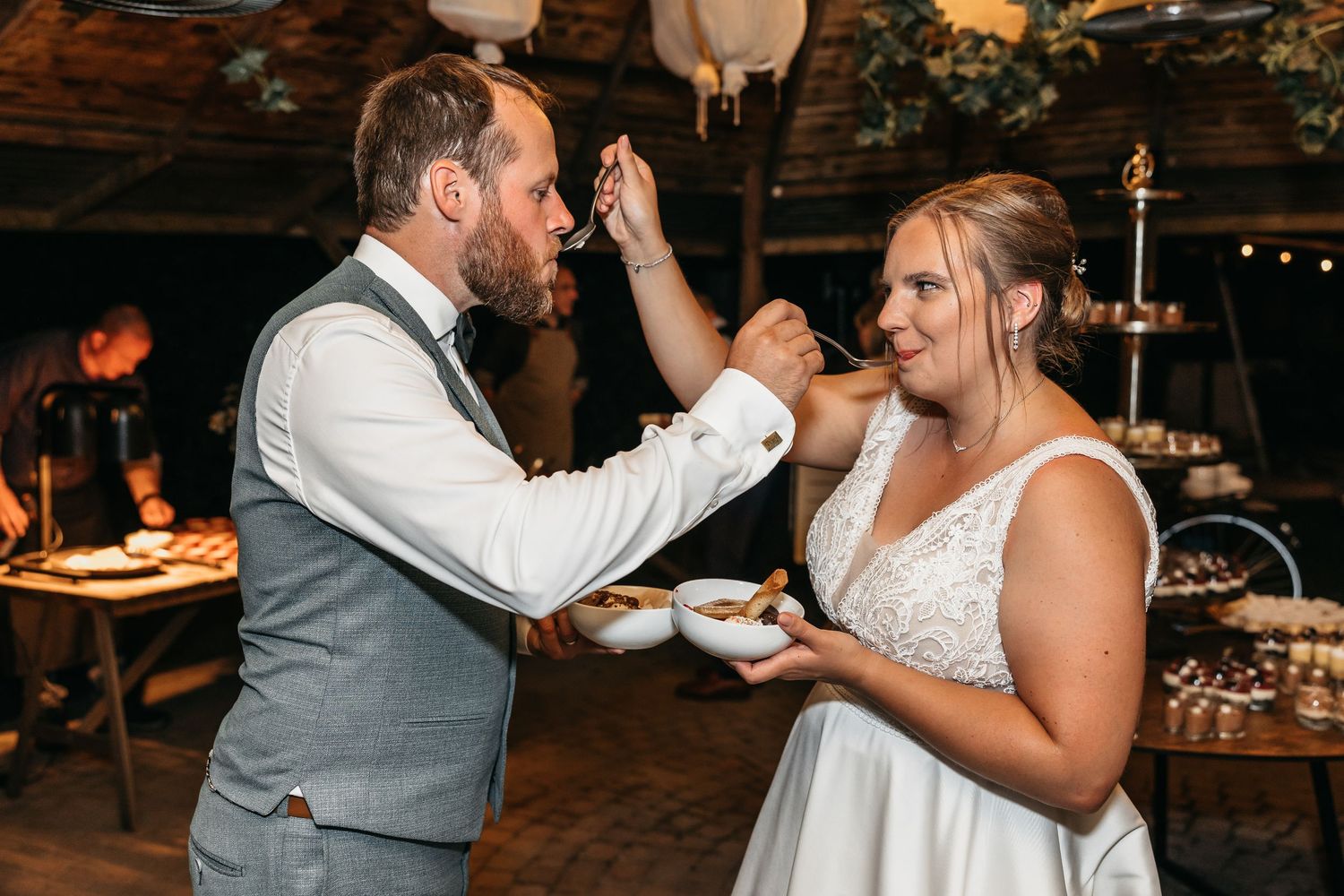 Bride and groom playfully feeding each other dessert at rustic wedding reception celebration.