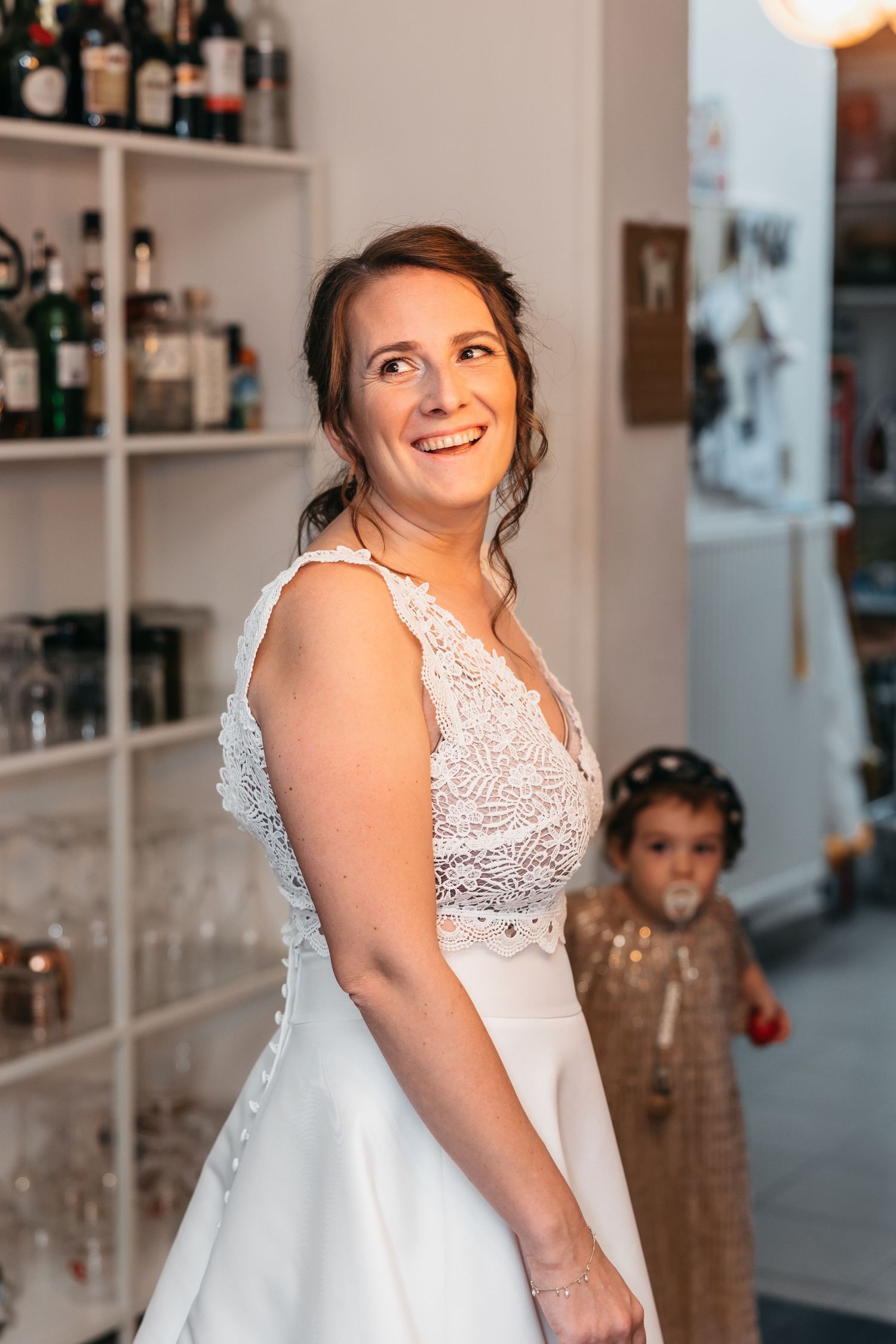 Smiling bride in white lace wedding dress with child in background at reception venue.