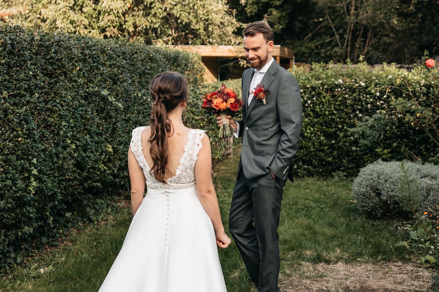 Groom in gray suit smiling at bride holding red and orange bouquet in sunny garden setting.