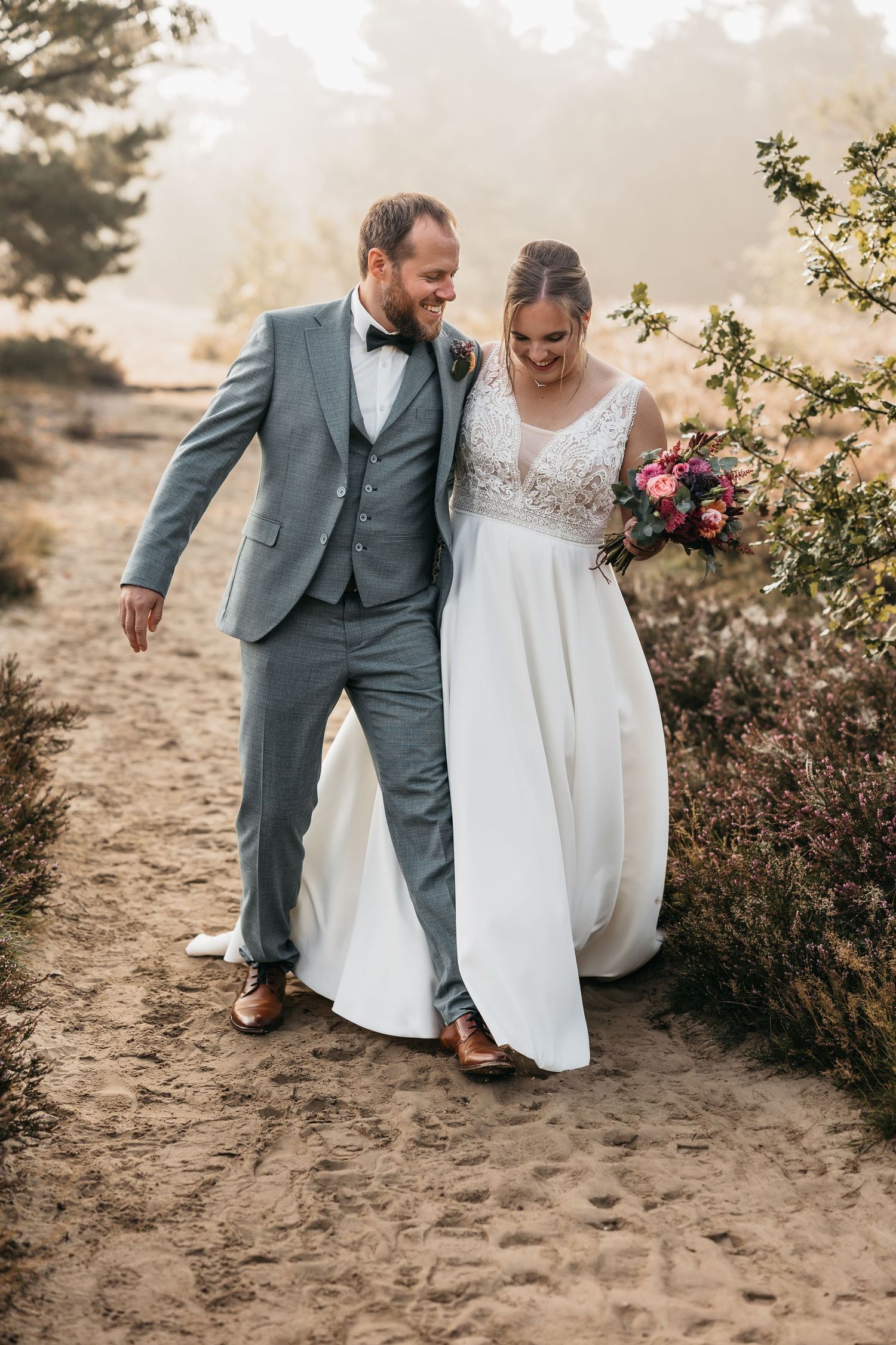 Laughing bride and groom walking along a sandy path through heathland, bride carrying a vibrant bouquet.