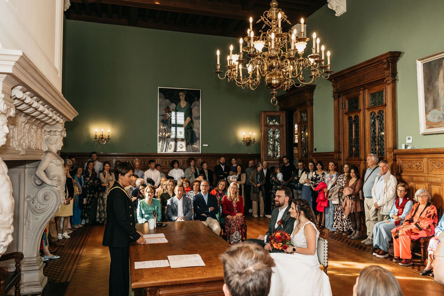 Wedding officiant reads vows as bride and groom stand at ceremonial table in elegant green-walled hall.