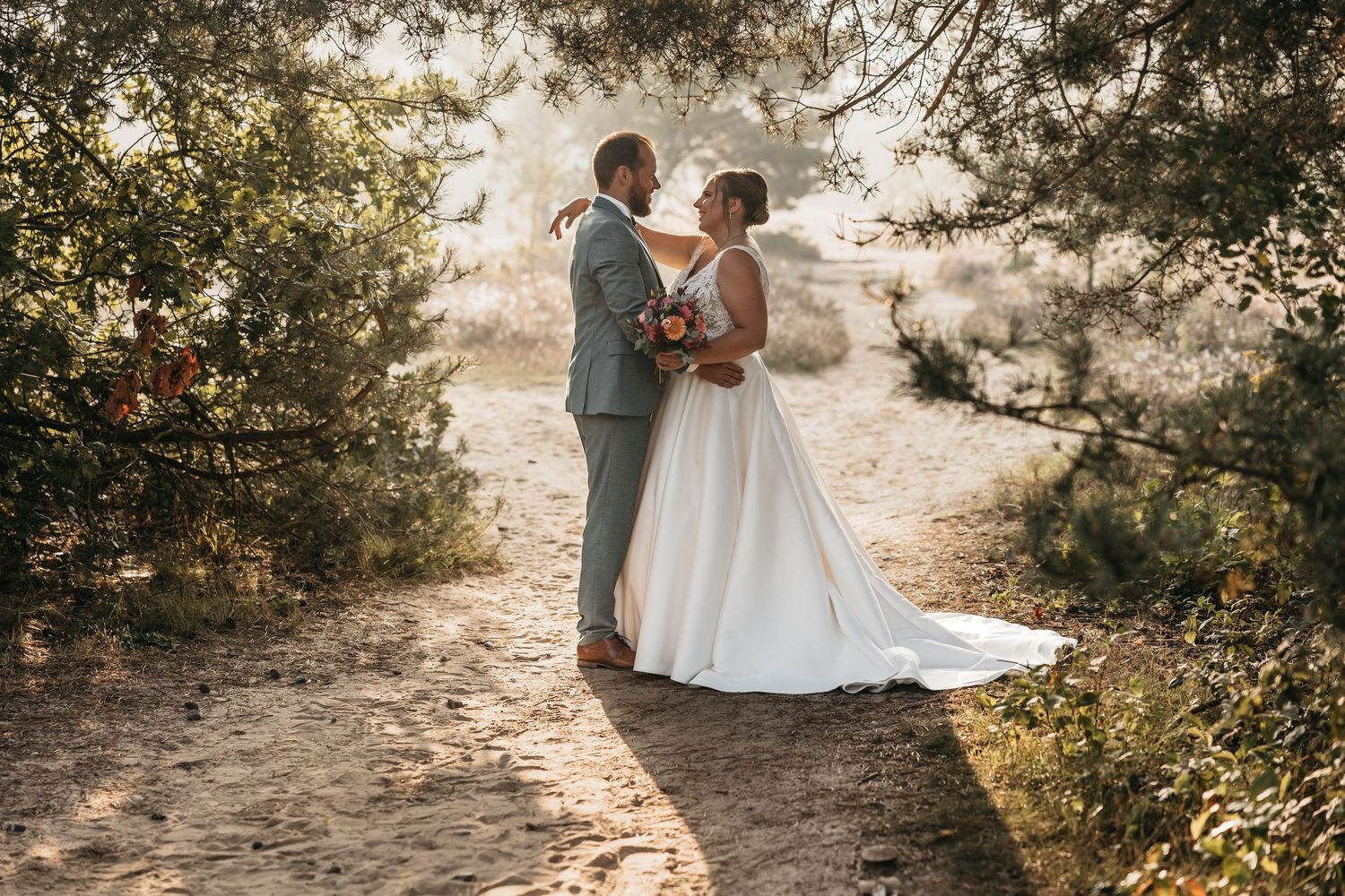 Wedding couple embraces tenderly on a sun-drenched sandy trail framed by pine trees in warm light.