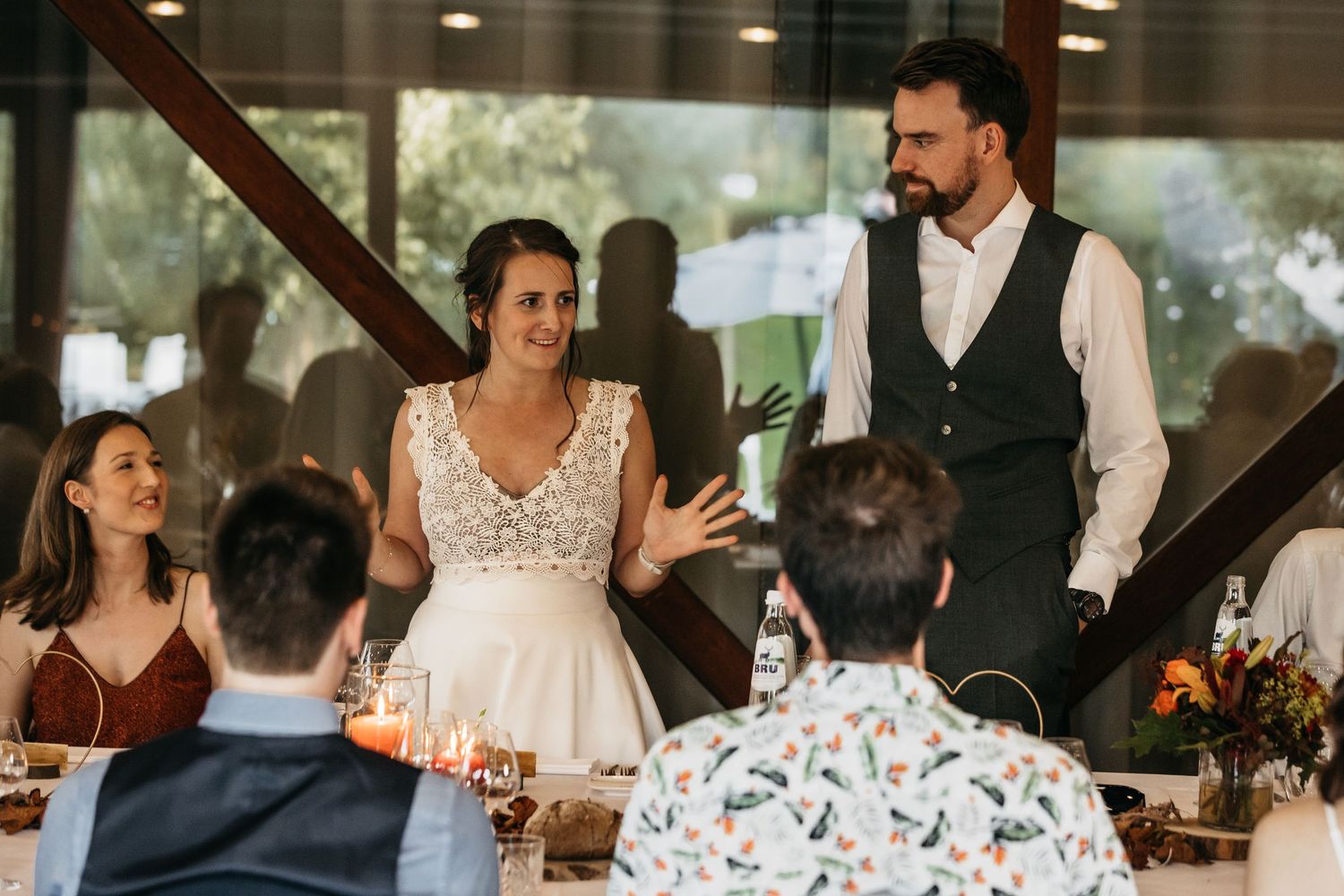 Bride and groom speaking to seated wedding guests at a candlelit reception dinner table.