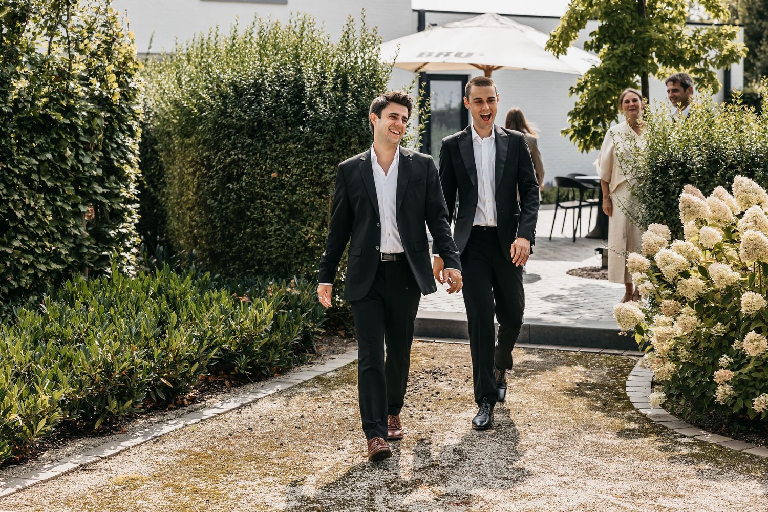 Two groomsmen in black suits walk hand in hand along a garden path at a sunny outdoor wedding.