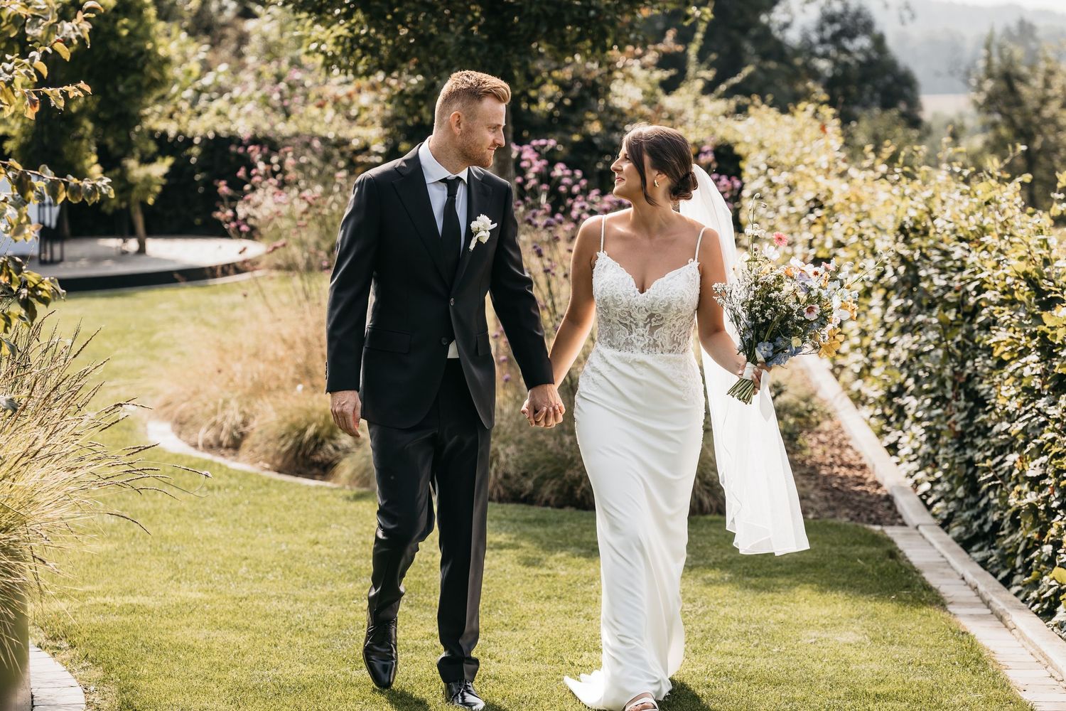 Newlyweds walking hand in hand through a beautiful garden, smiling at each other on their wedding day.