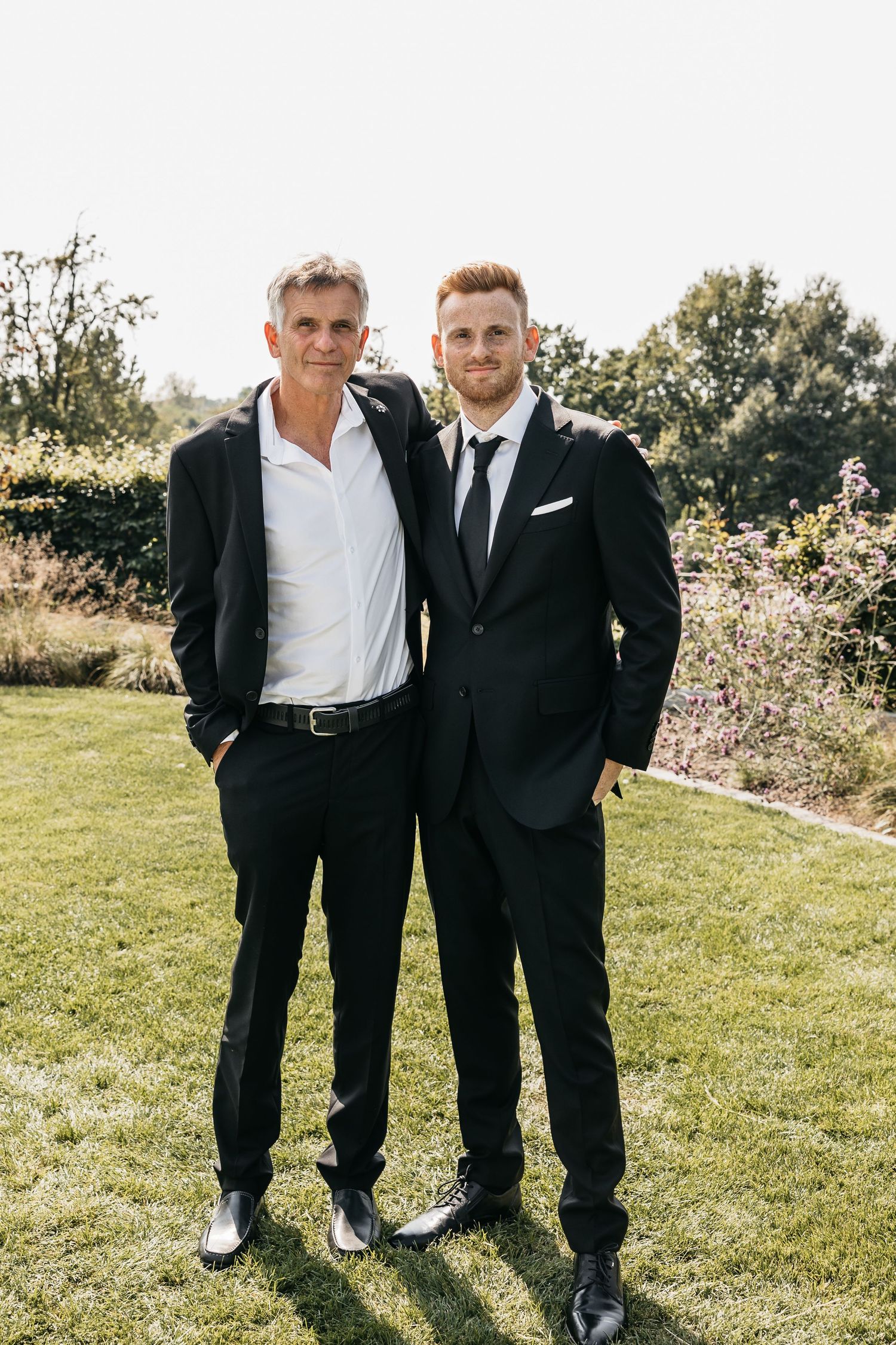Two men in black suits pose together on a green lawn at an outdoor wedding celebration.