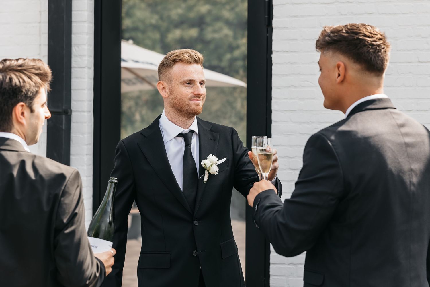 Three groomsmen in black suits toast with champagne glasses outdoors before a wedding.
