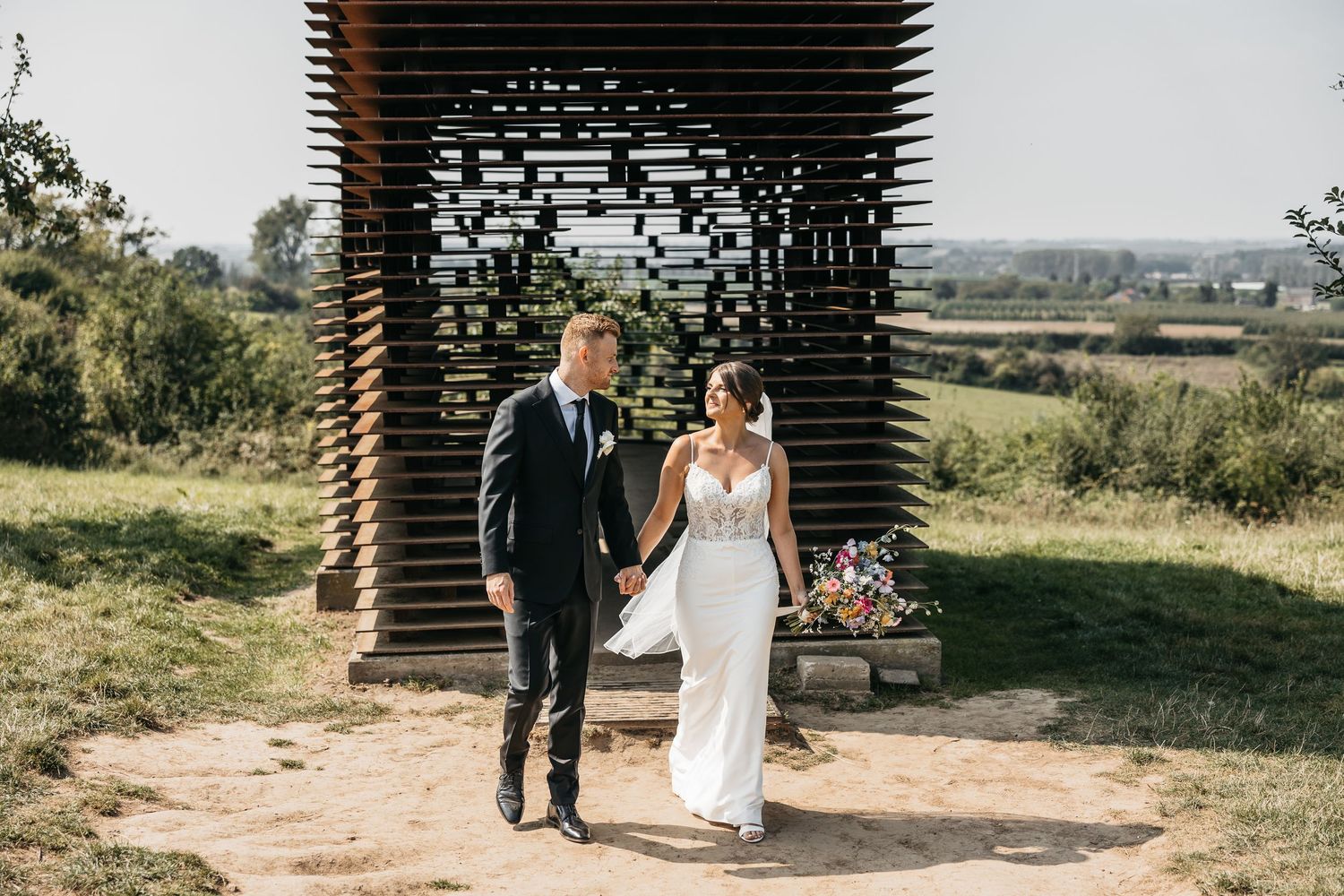Bride and groom walk together holding hands with a modern wooden lattice structure in the background.
