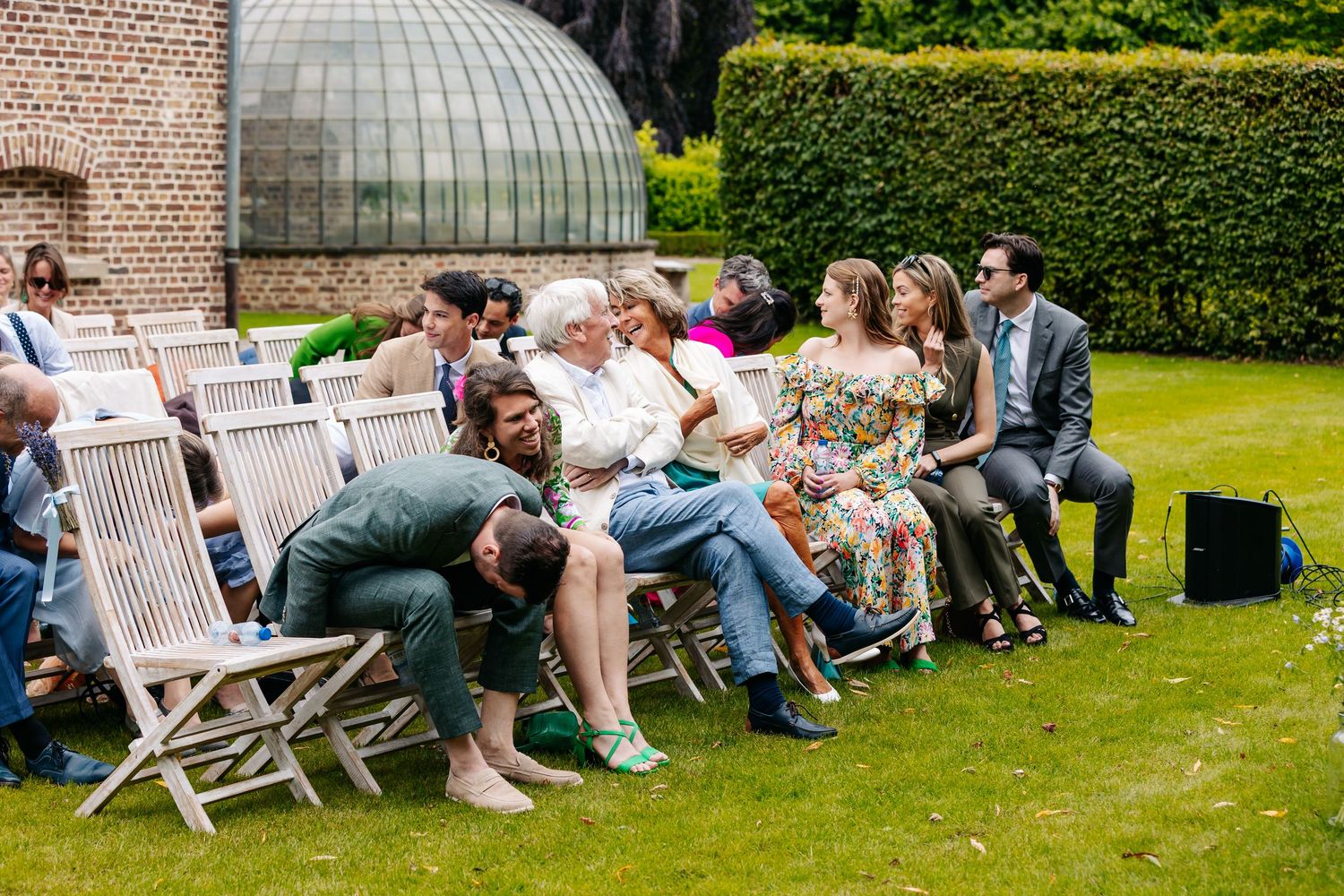 Wedding guests relaxing and laughing on wooden chairs during an outdoor garden ceremony on a sunny day.