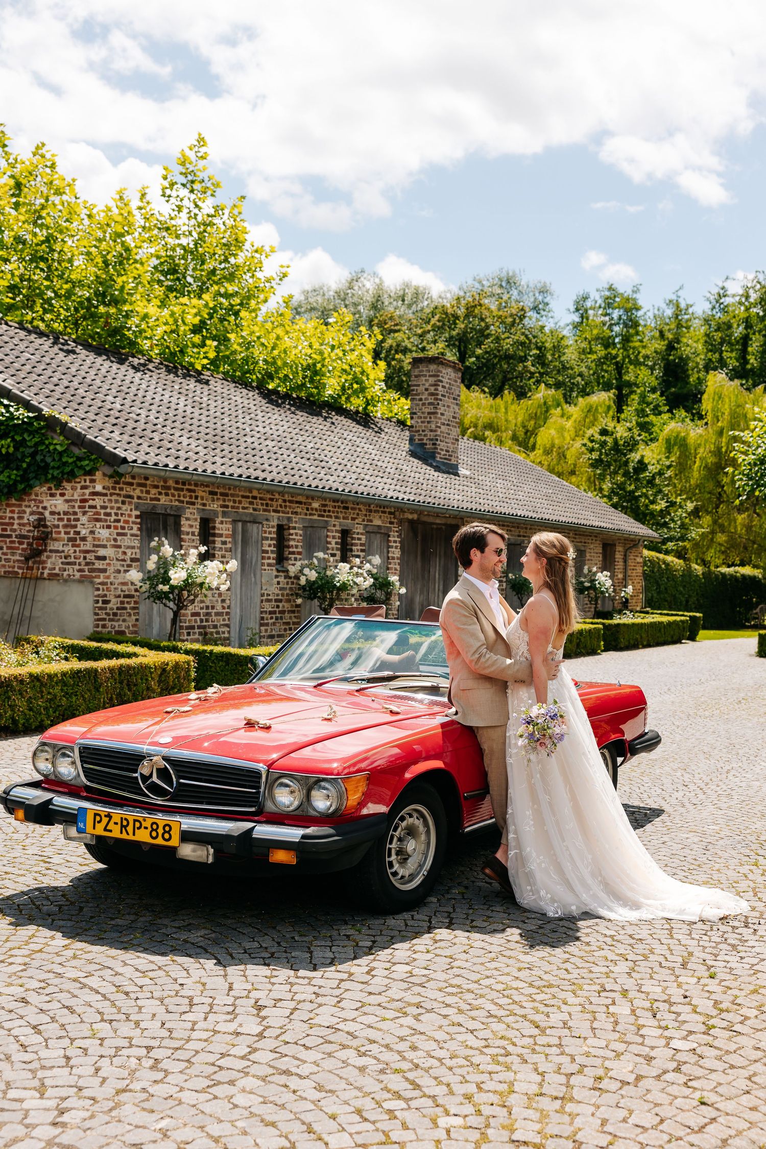 Bride and groom embrace beside a classic red Mercedes convertible on cobblestones.