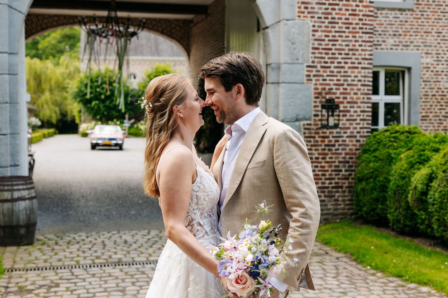 Bride and groom sharing romantic moment outdoors at brick manor, groom in tan suit holding bouquet.