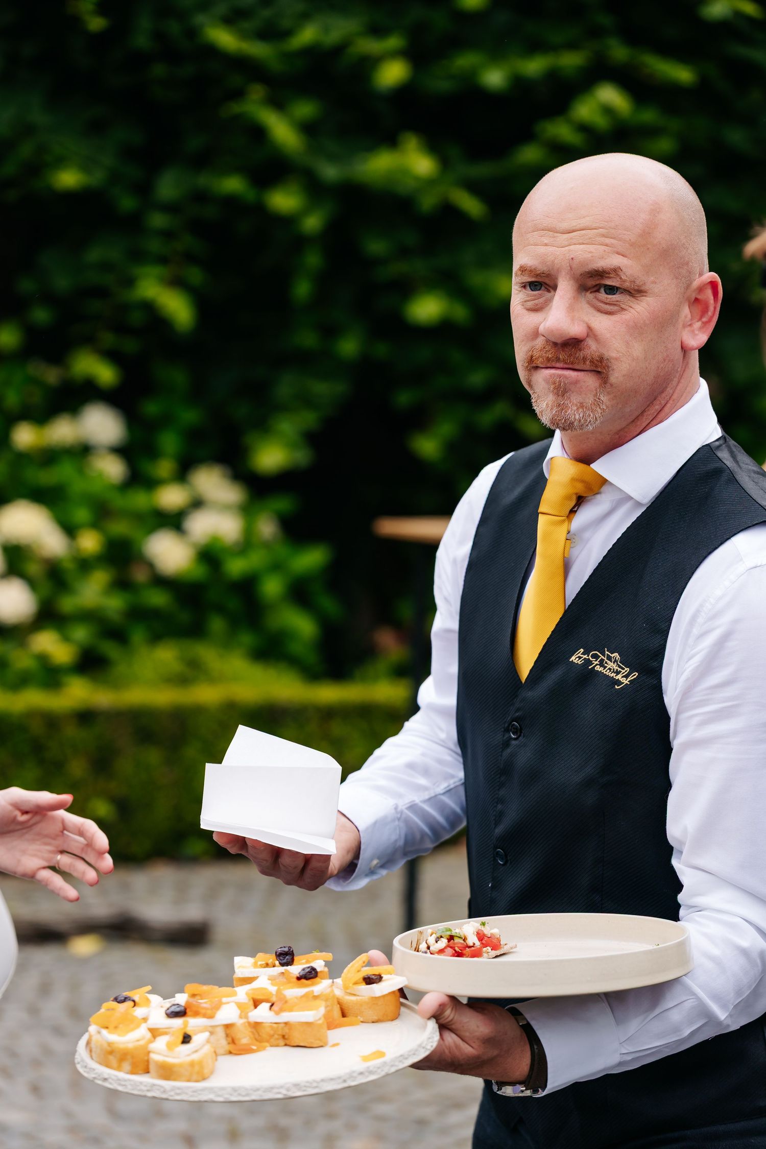 Bald waiter in vest and yellow tie serving elegant canapes on trays at outdoor event.