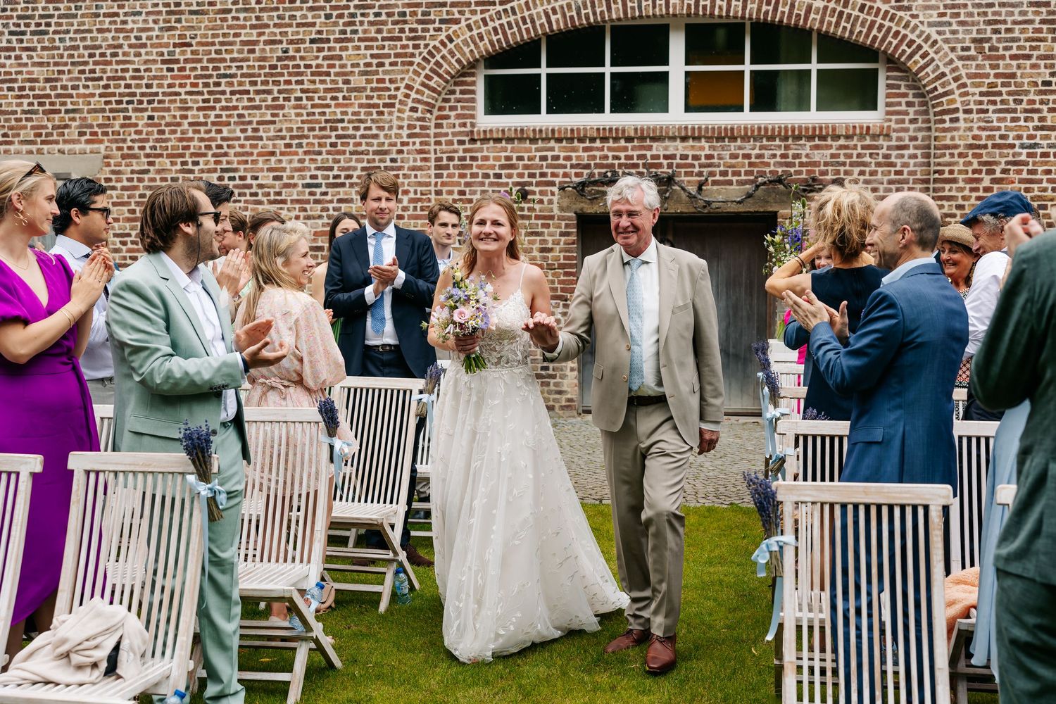 Bride walks down the aisle with her father at an outdoor wedding ceremony beside a brick building.