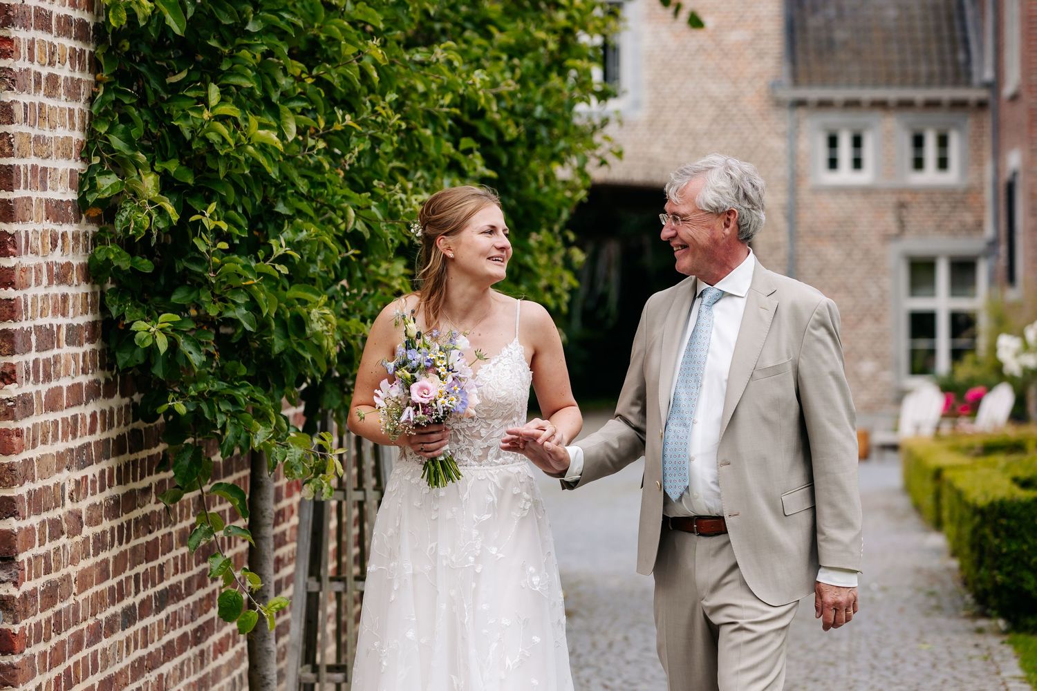 Bride in lace gown walking with older gentleman along brick wall with bouquet.