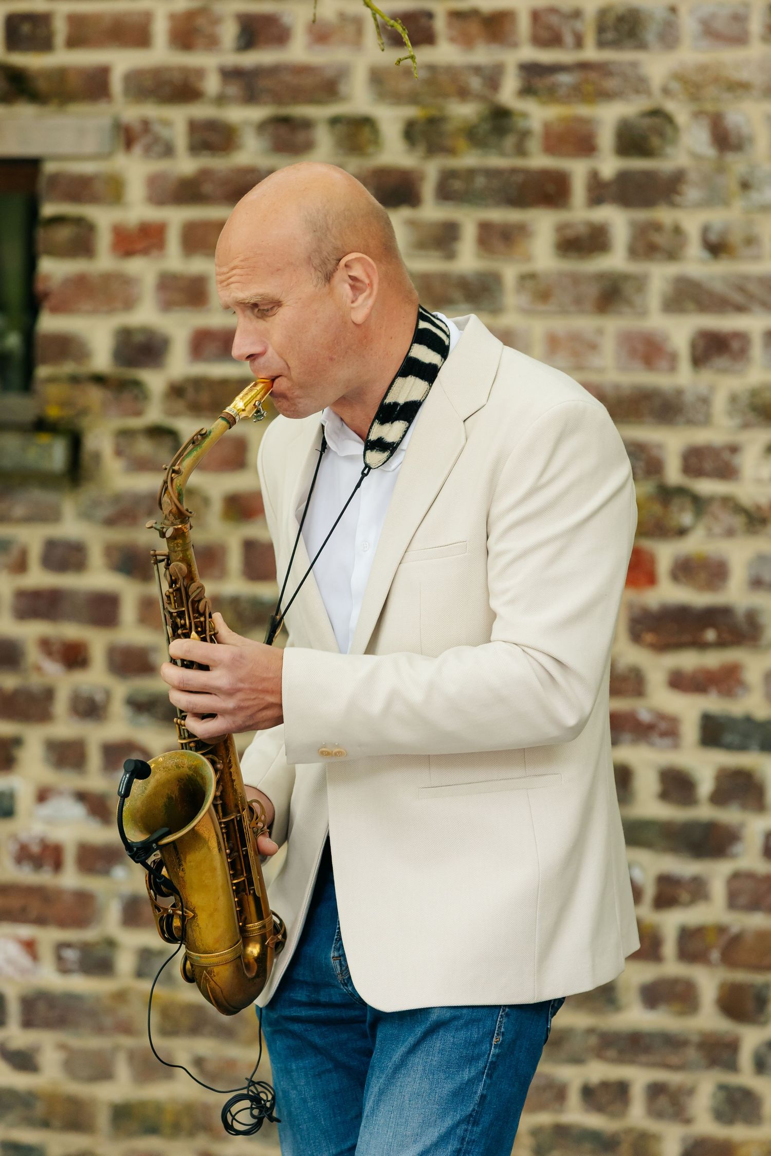 Bald musician in a white blazer playing a saxophone against a rustic brick wall backdrop.