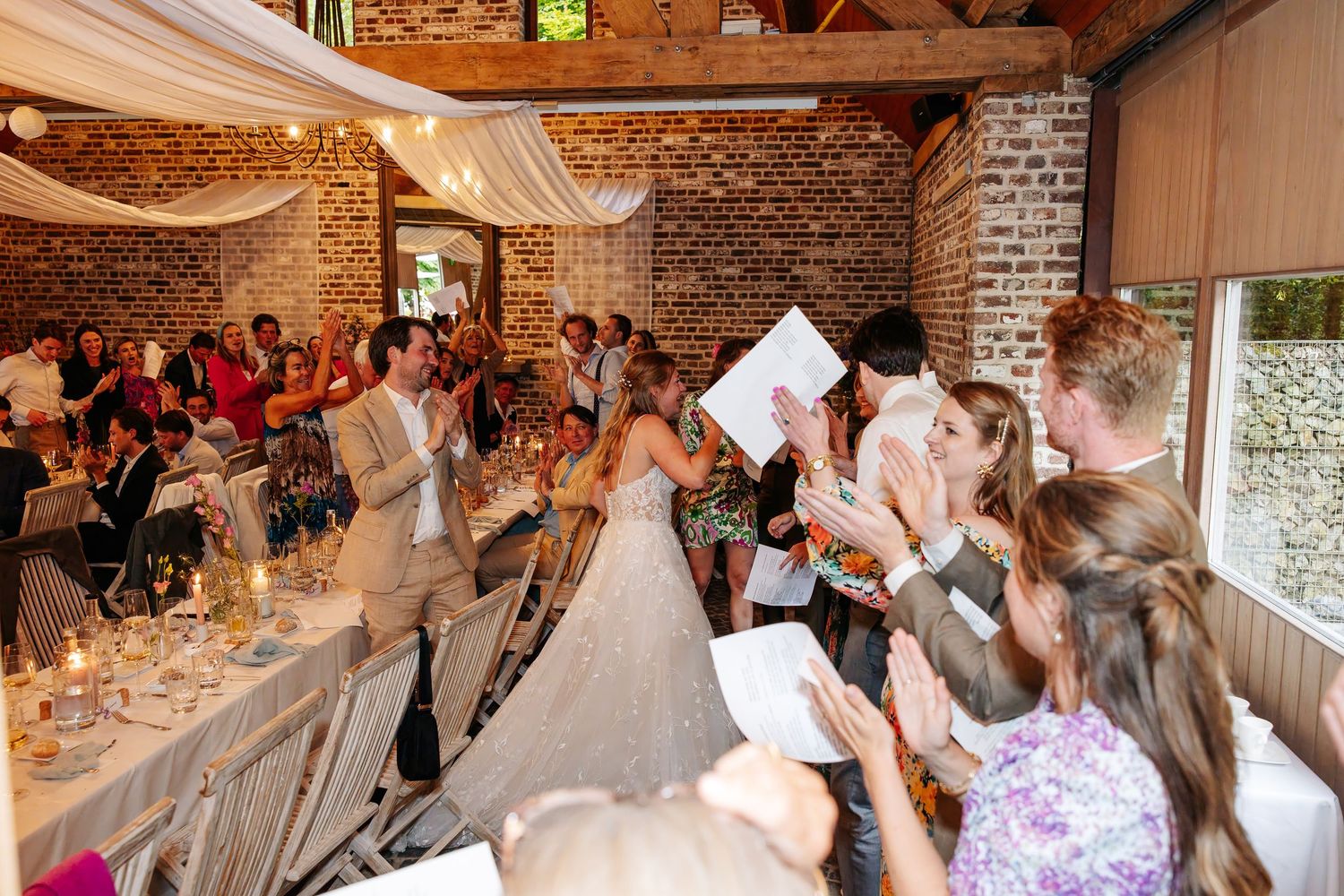 Bride and groom entering reception hall as guests cheer and applaud in rustic venue.