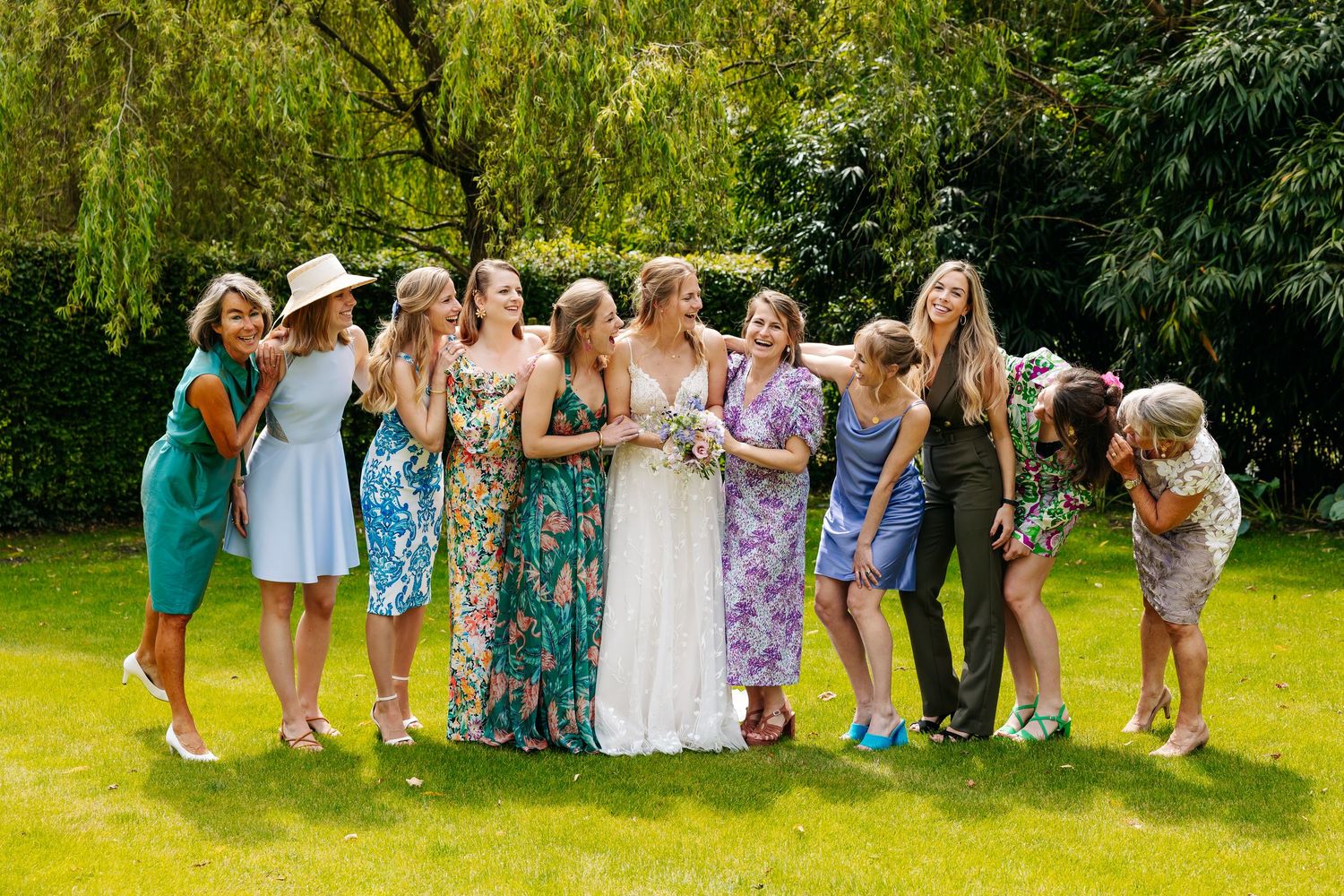 Bride in white dress surrounded by bridesmaids in colorful floral dresses laughing on green lawn.
