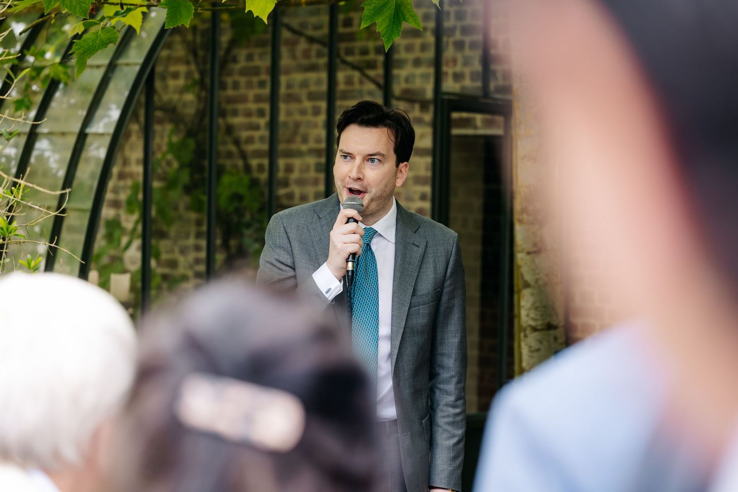 Elegant man in grey suit speaks into microphone at outdoor wedding reception near brick wall.