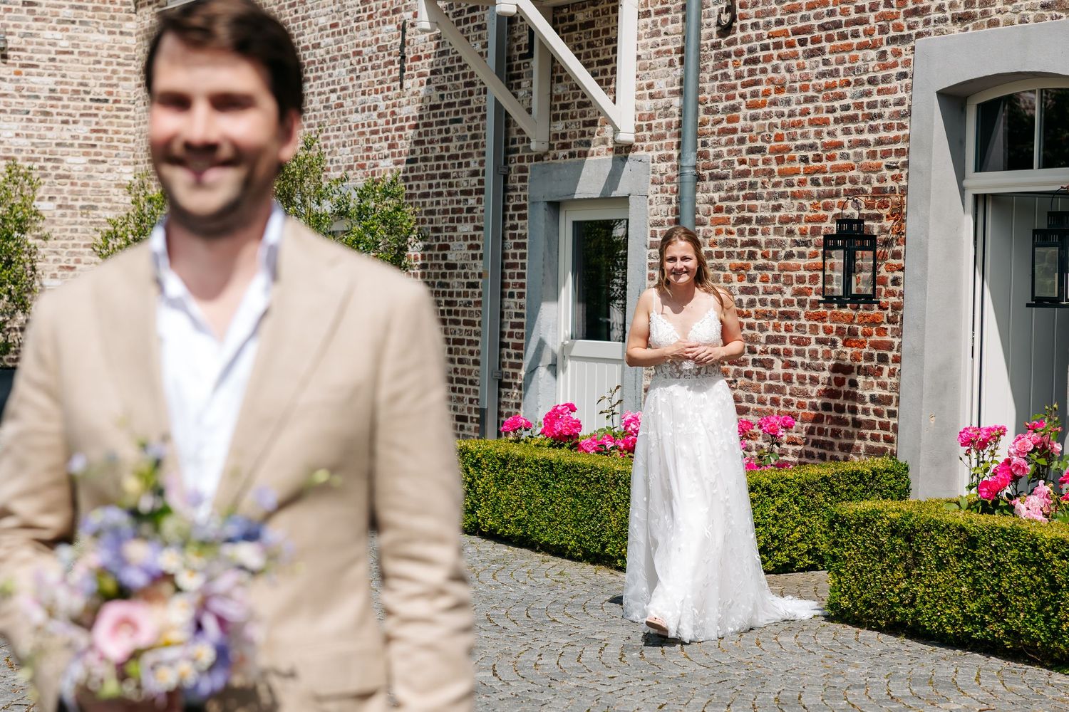 Bride in white gown smiles at groom holding bouquet during outdoor first look at brick estate venue.