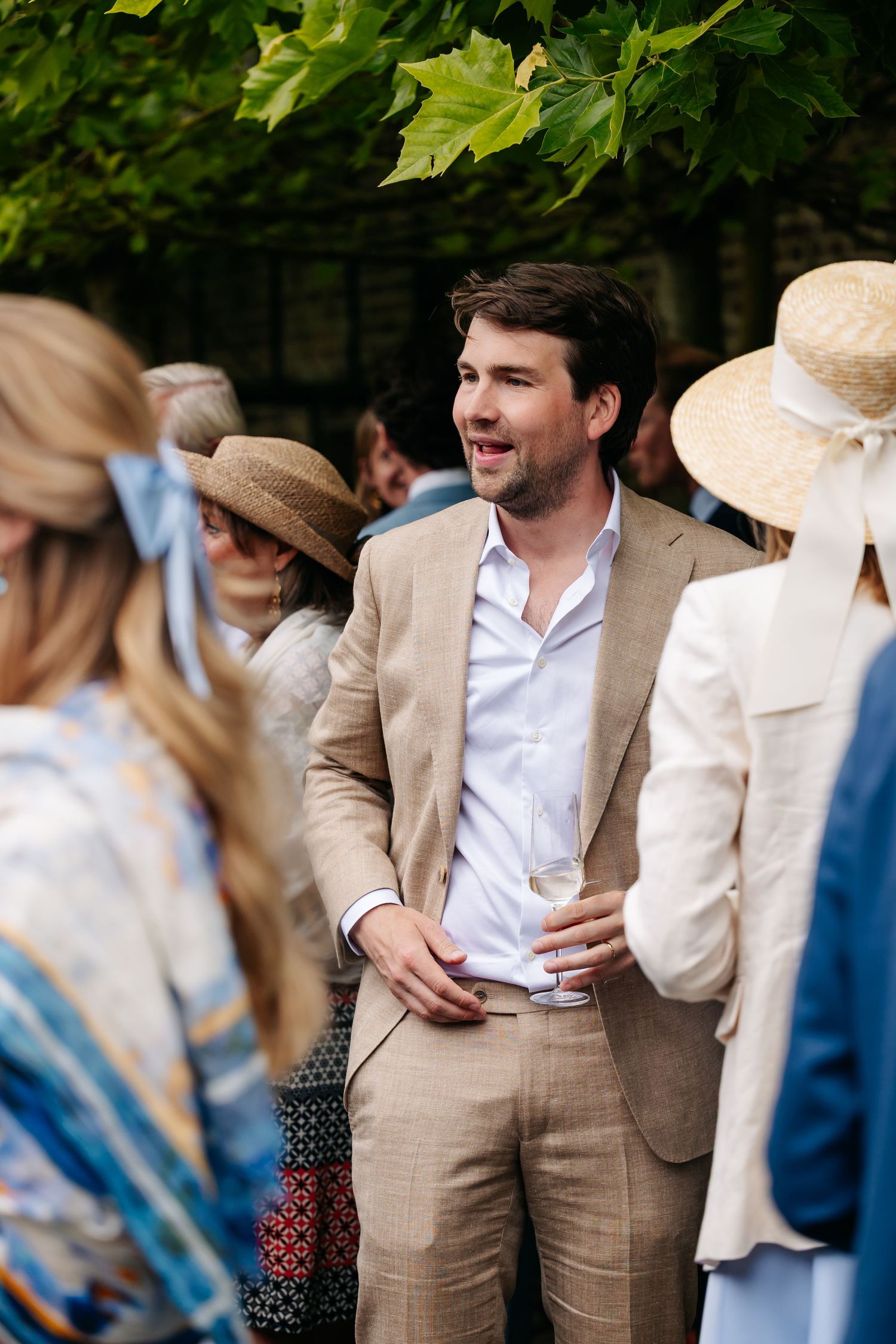 A man in a beige linen suit holds a drink and mingles at an outdoor summer wedding.