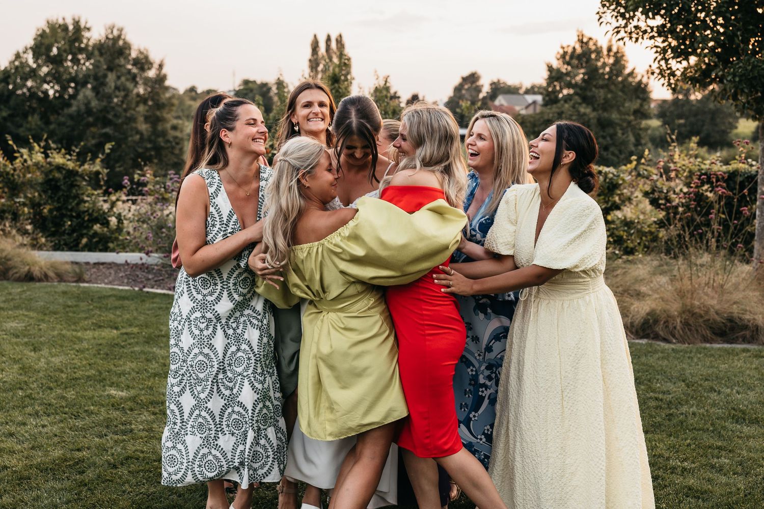 Small group of female wedding guests laughing and hugging in garden at golden hour.
