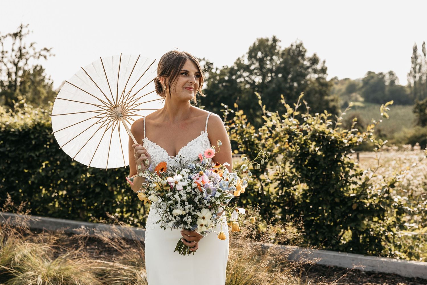 Elegant bride in white gown holding a colorful wildflower bouquet and white parasol in a garden setting.